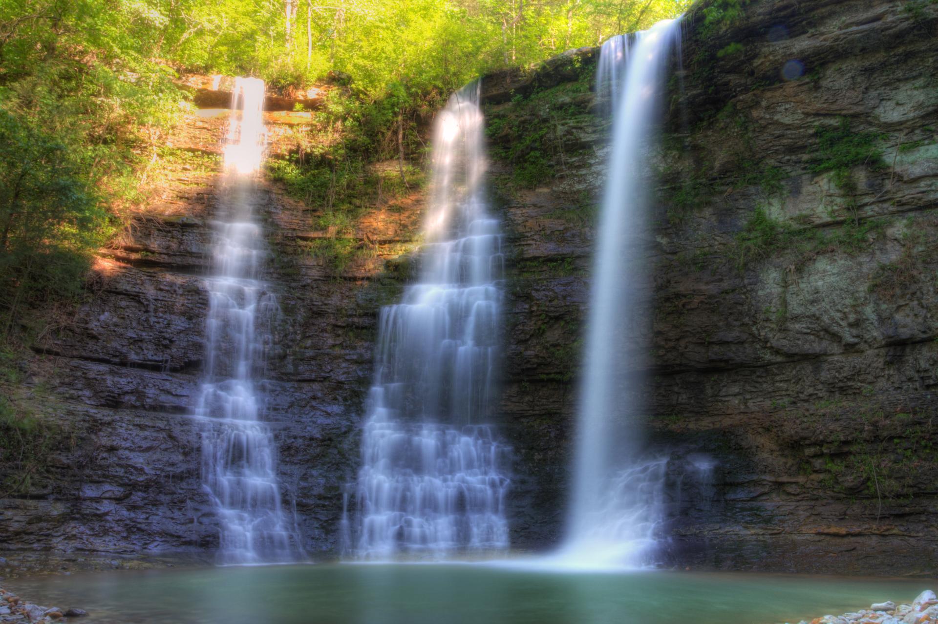Three waterfalls cascade down rocky cliffs into a serene pool, bathed in sunlight.