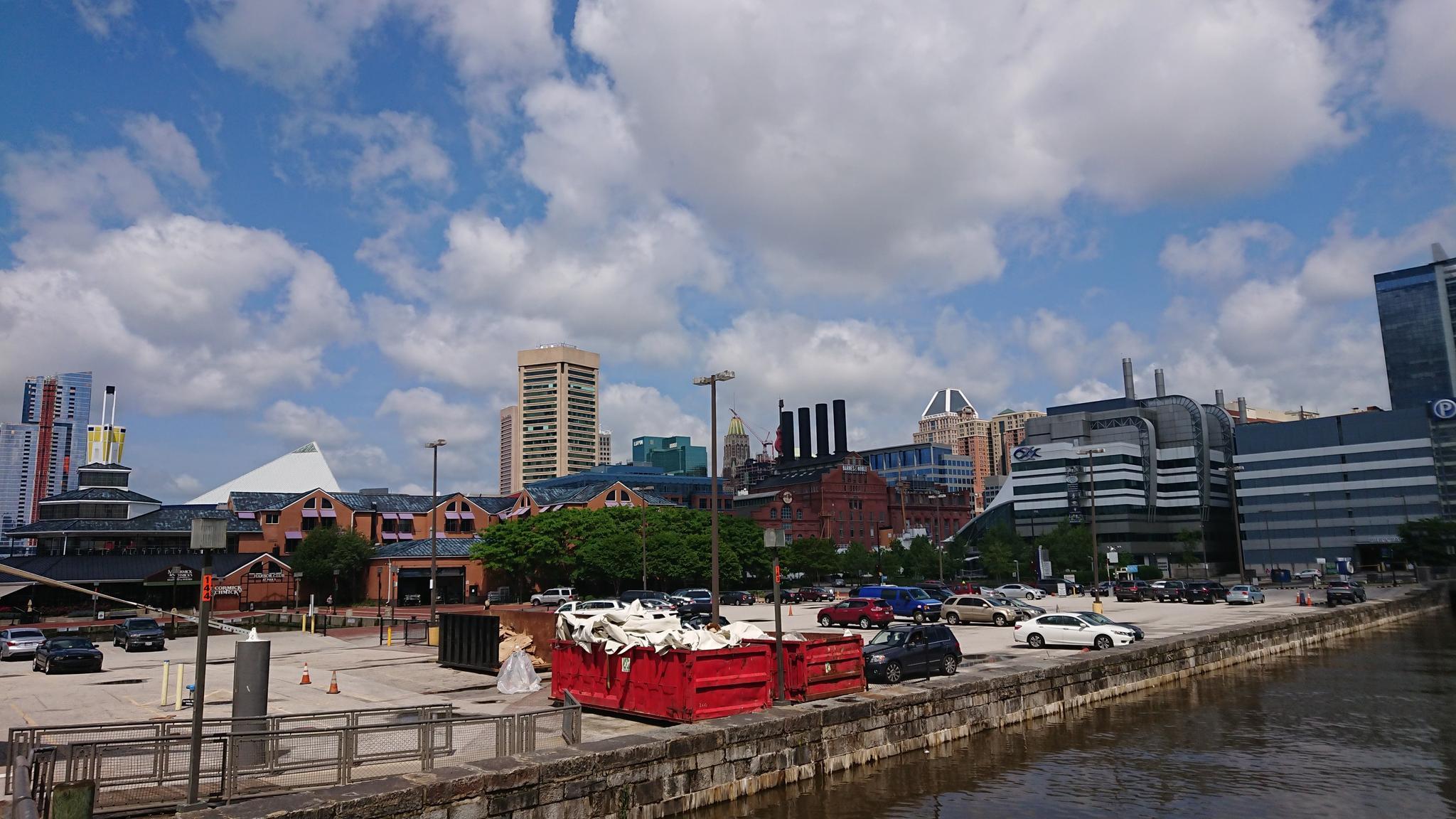 A red dumpster on a paved waterfront, with city buildings and a cloudy sky.