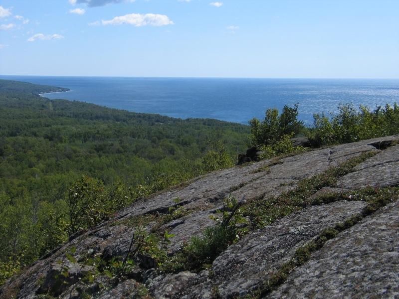 Sunny view from a rocky cliff over a green forest and blue water.