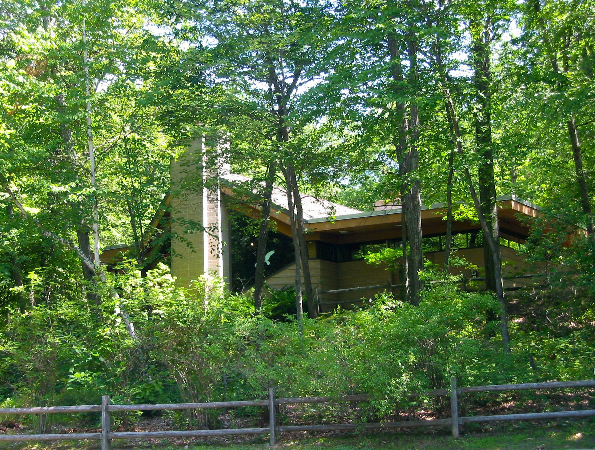 Modern house partially hidden by dense green trees, seen through a fence.