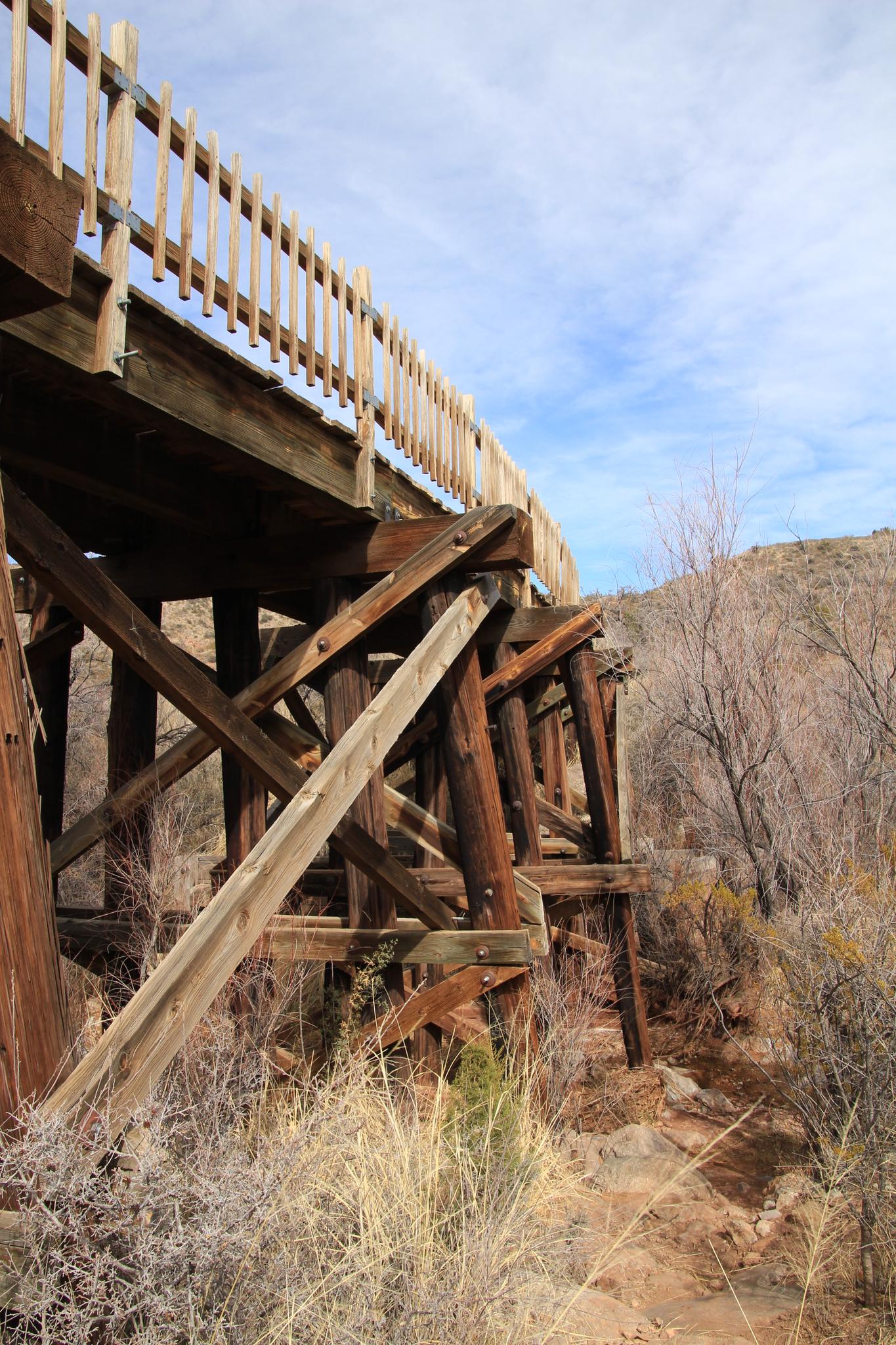 A wooden trestle bridge stands over dry brush under a partly cloudy sky.