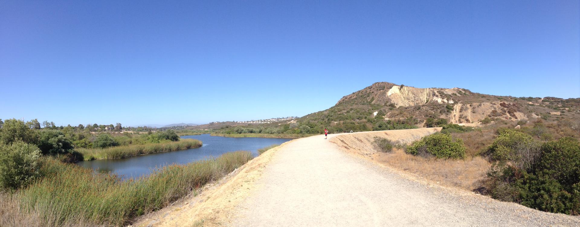 A wide dirt path alongside a lake, with a large hill under a clear sky.