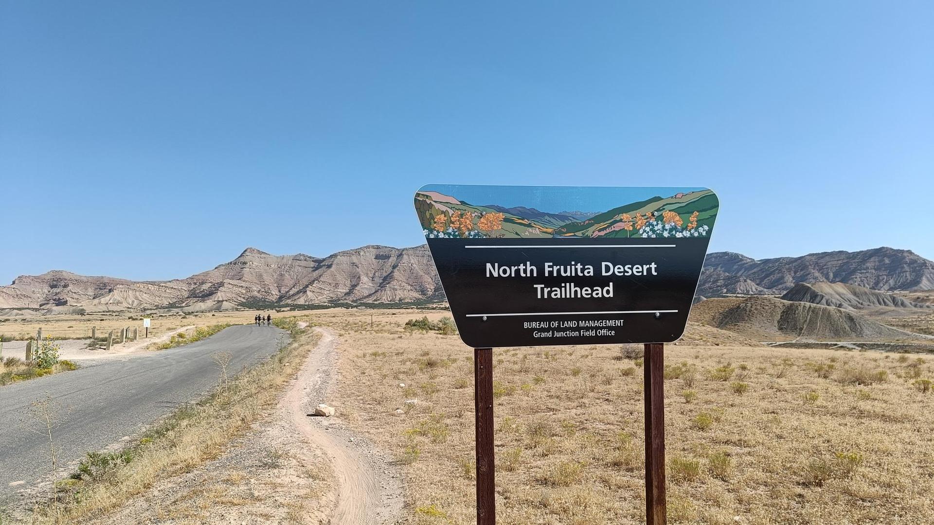 Trailhead sign in a dry, mountainous landscape with a road under clear sky.