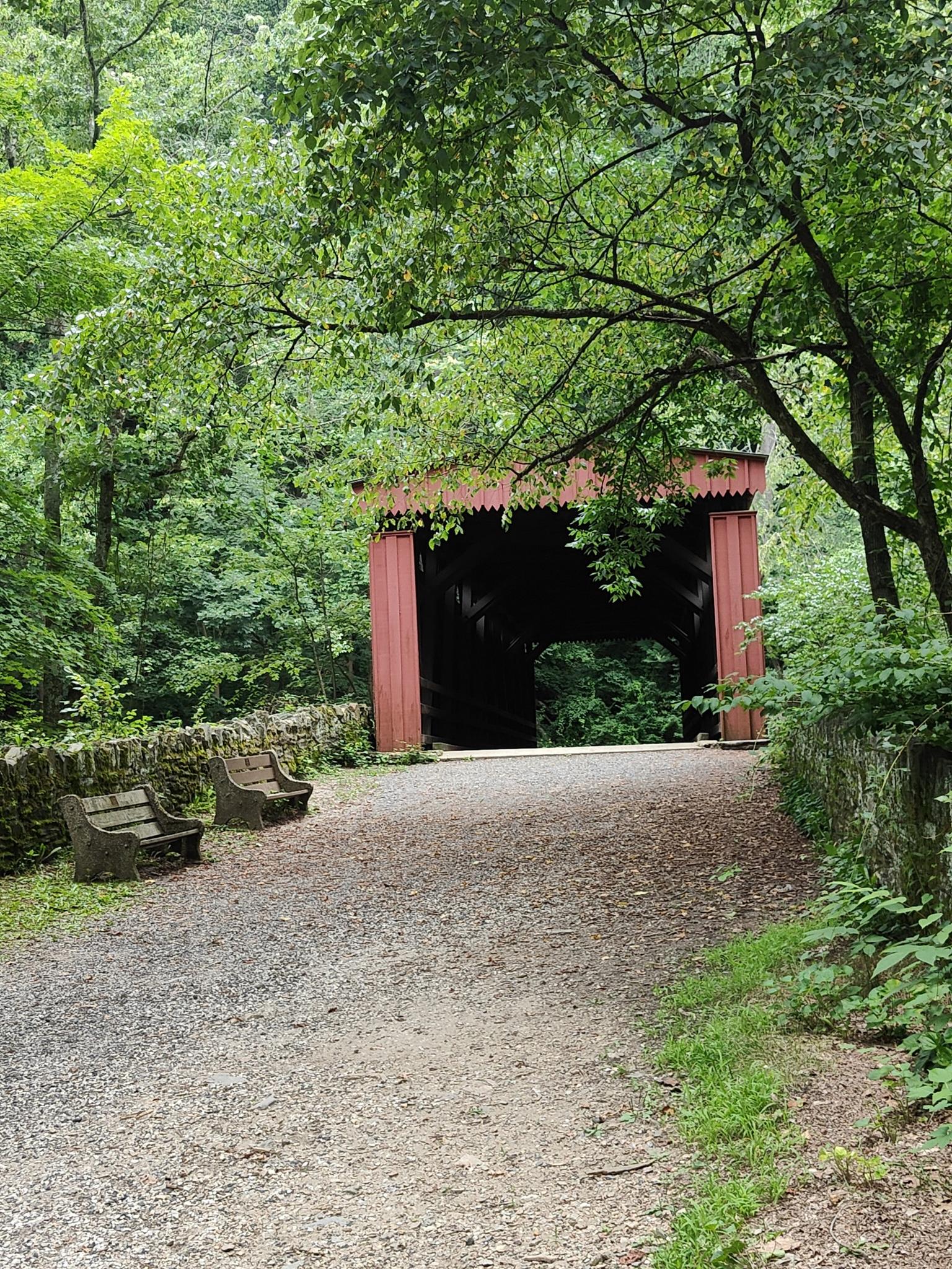 A gravel path leads to a red covered bridge surrounded by green trees.