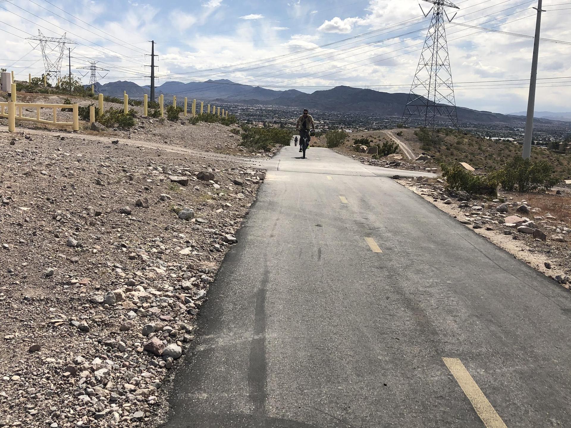 Cyclist on a paved path in a desert landscape with power lines and mountains.