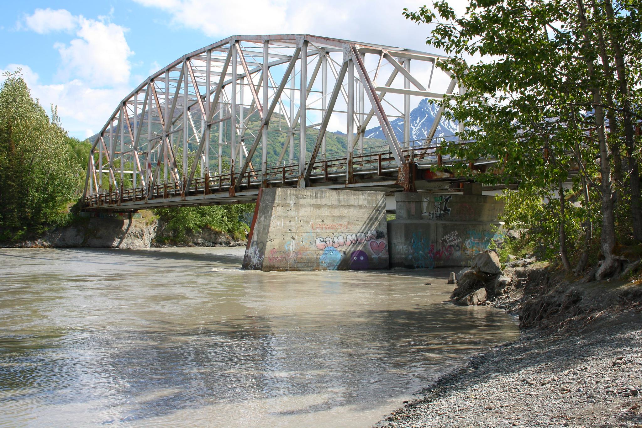 Large metal truss bridge spans a muddy river with mountains in background.