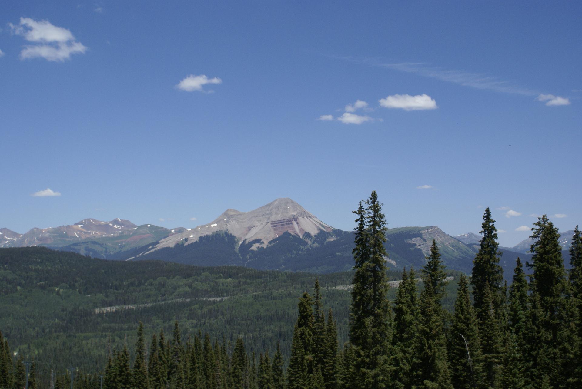 Majestic mountain peak rises above a dense evergreen forest under a bright blue sky.