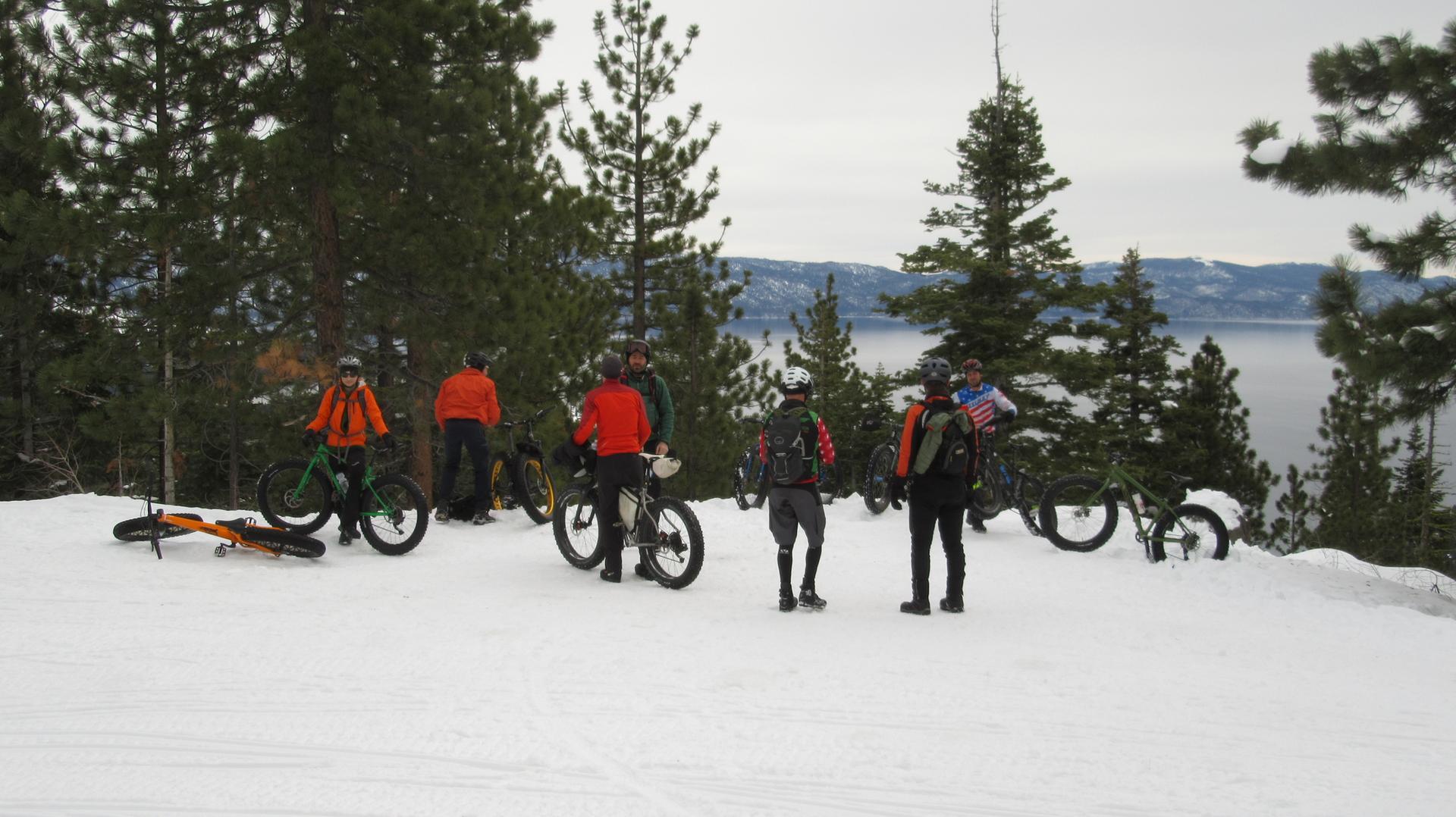 Group of fat bikers resting on a snowy hill overlooking a lake.