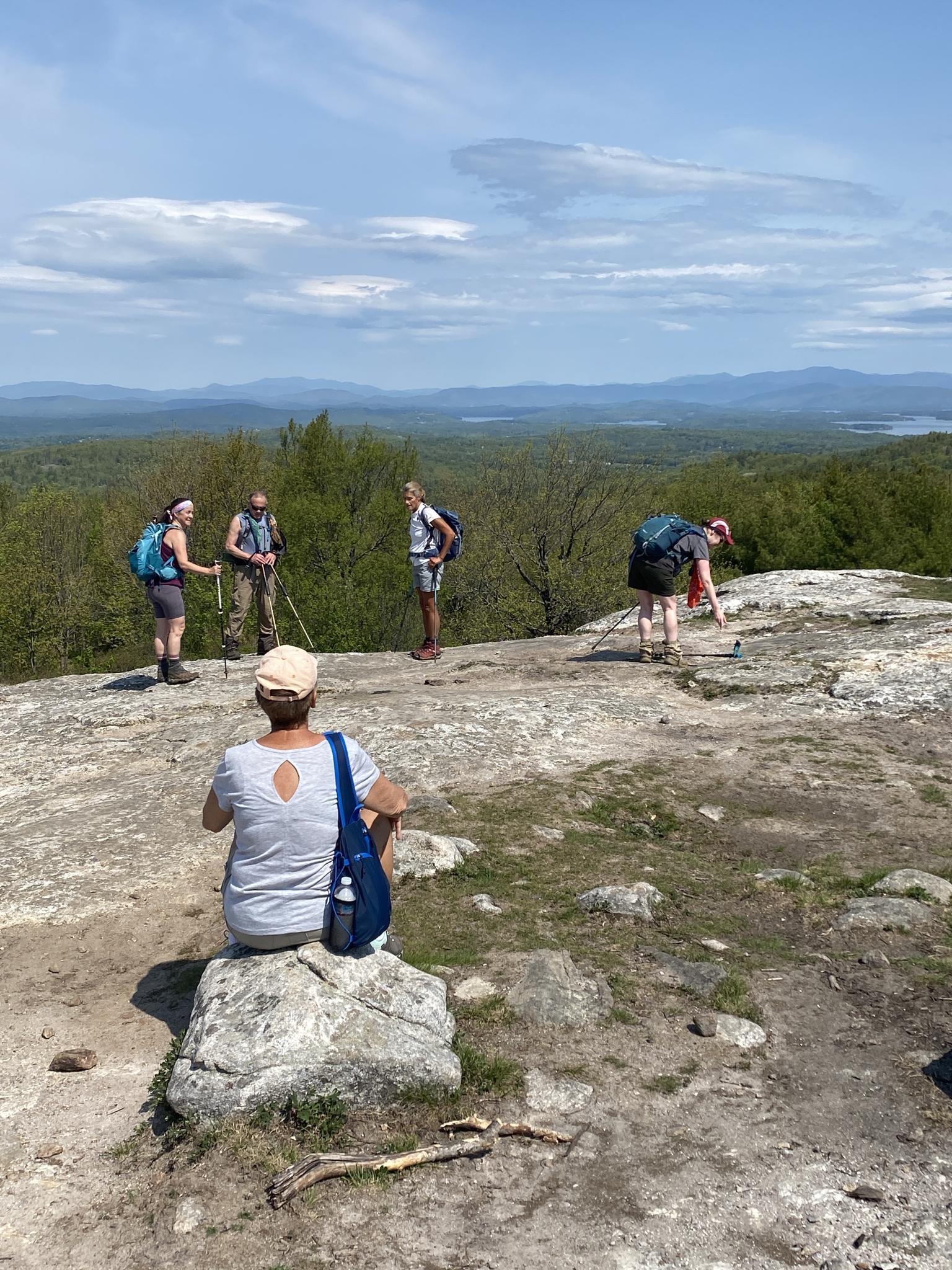 Hikers on a sunny, rocky summit overlooking a vast forested landscape.