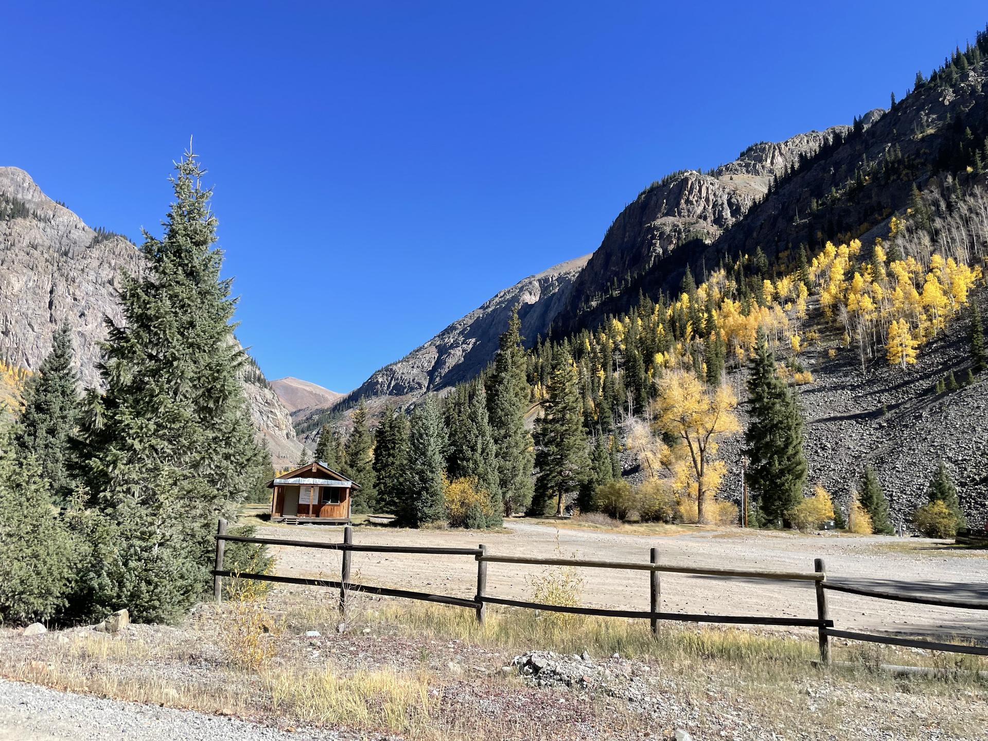 A small cabin sits in a mountain valley with bright autumn trees.