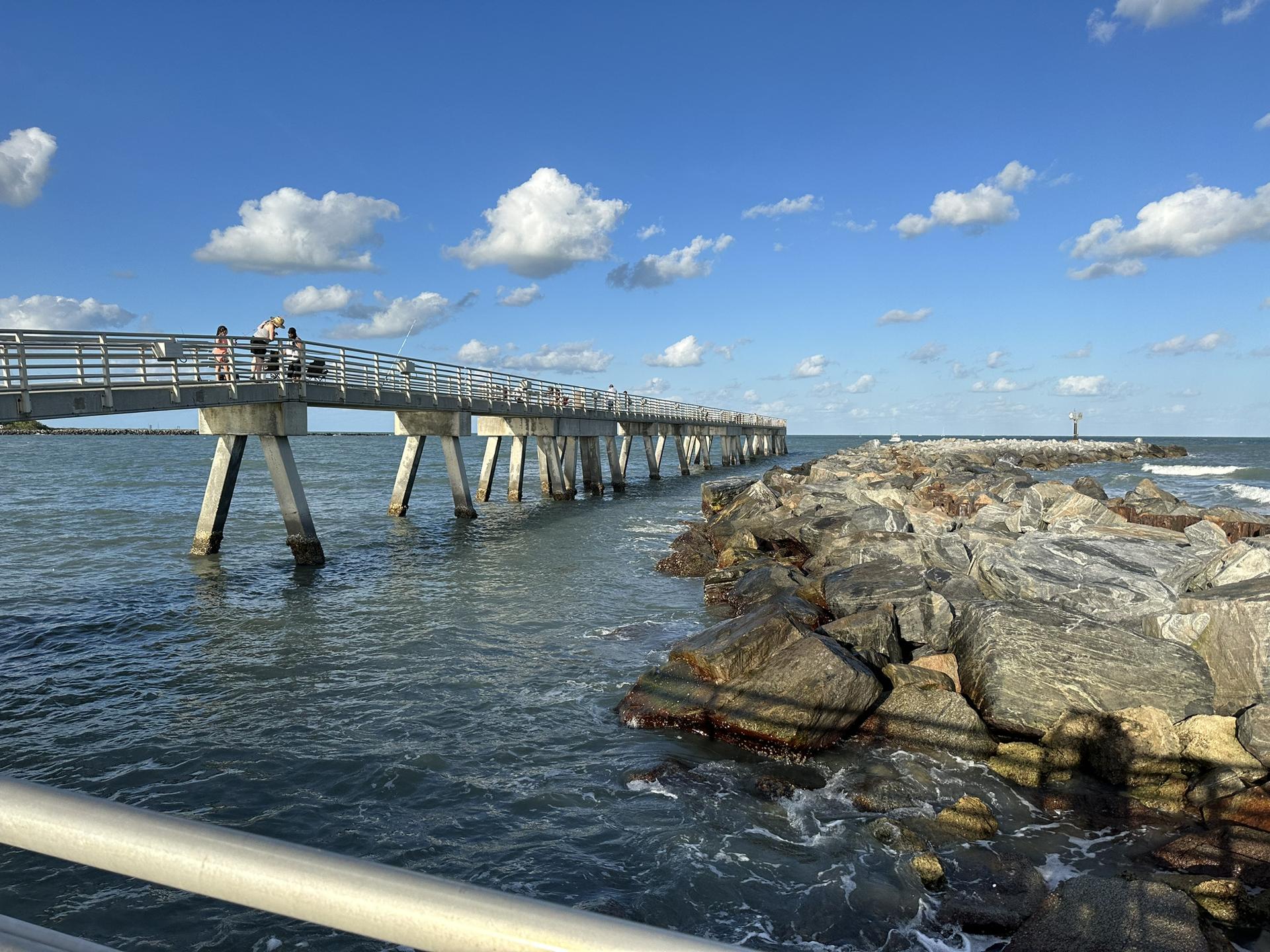 Sunny day with people on a long pier next to a rocky jetty.