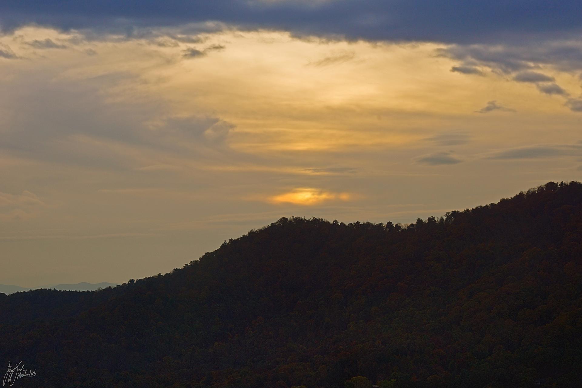 A vibrant sunset over a dark, forested mountain ridge with dramatic clouds.