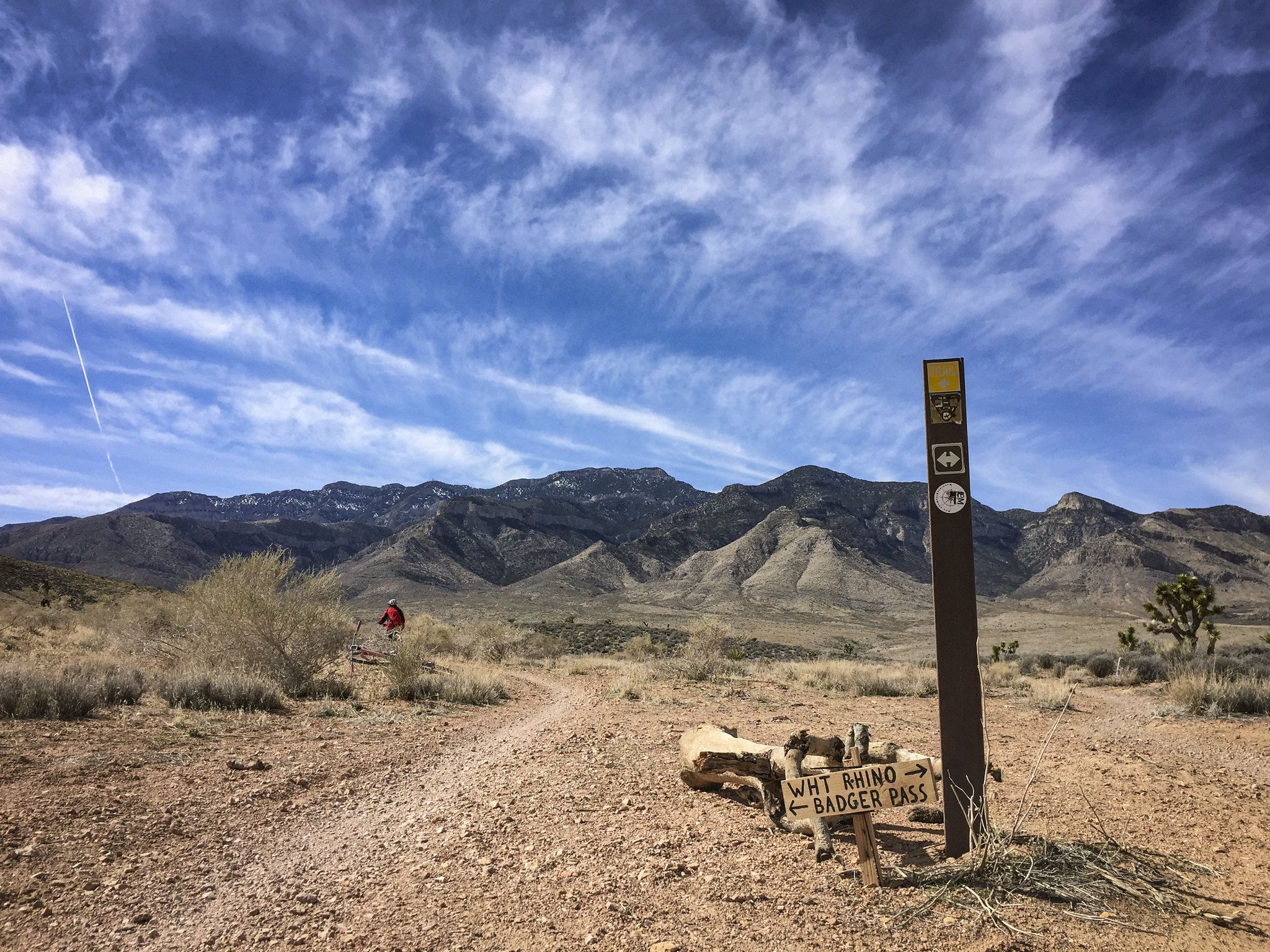 A trail sign in a desert landscape with mountains under a cloudy blue sky. - Badger Pass Loop