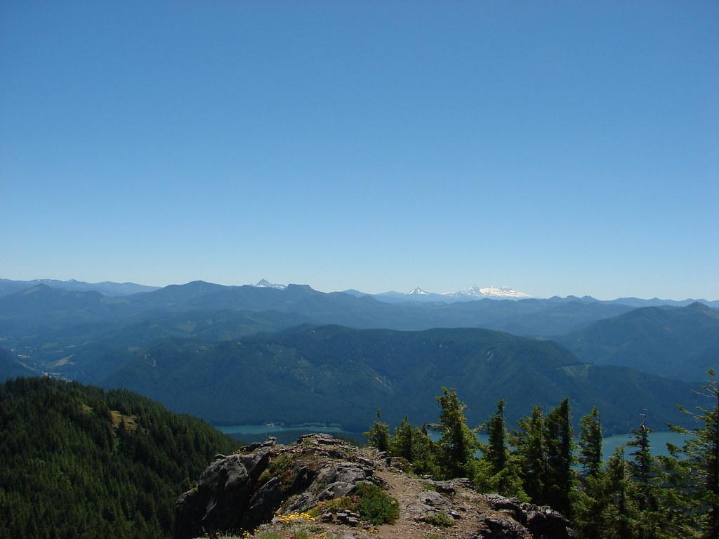 Sunny mountain vista with forested slopes, a lake, and distant snow-capped peaks.
