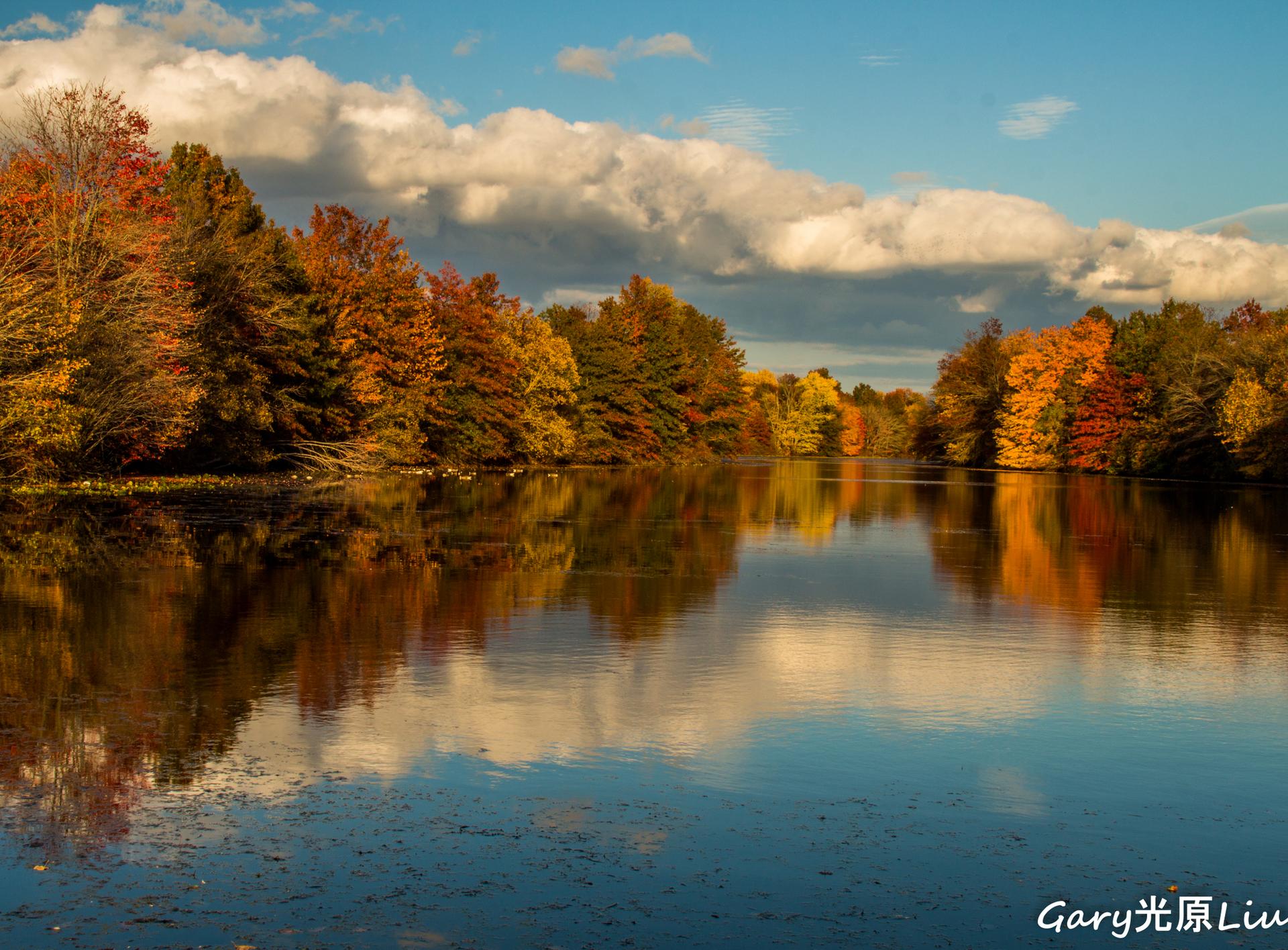A serene lake reflects colorful autumn trees under a partly cloudy blue sky.