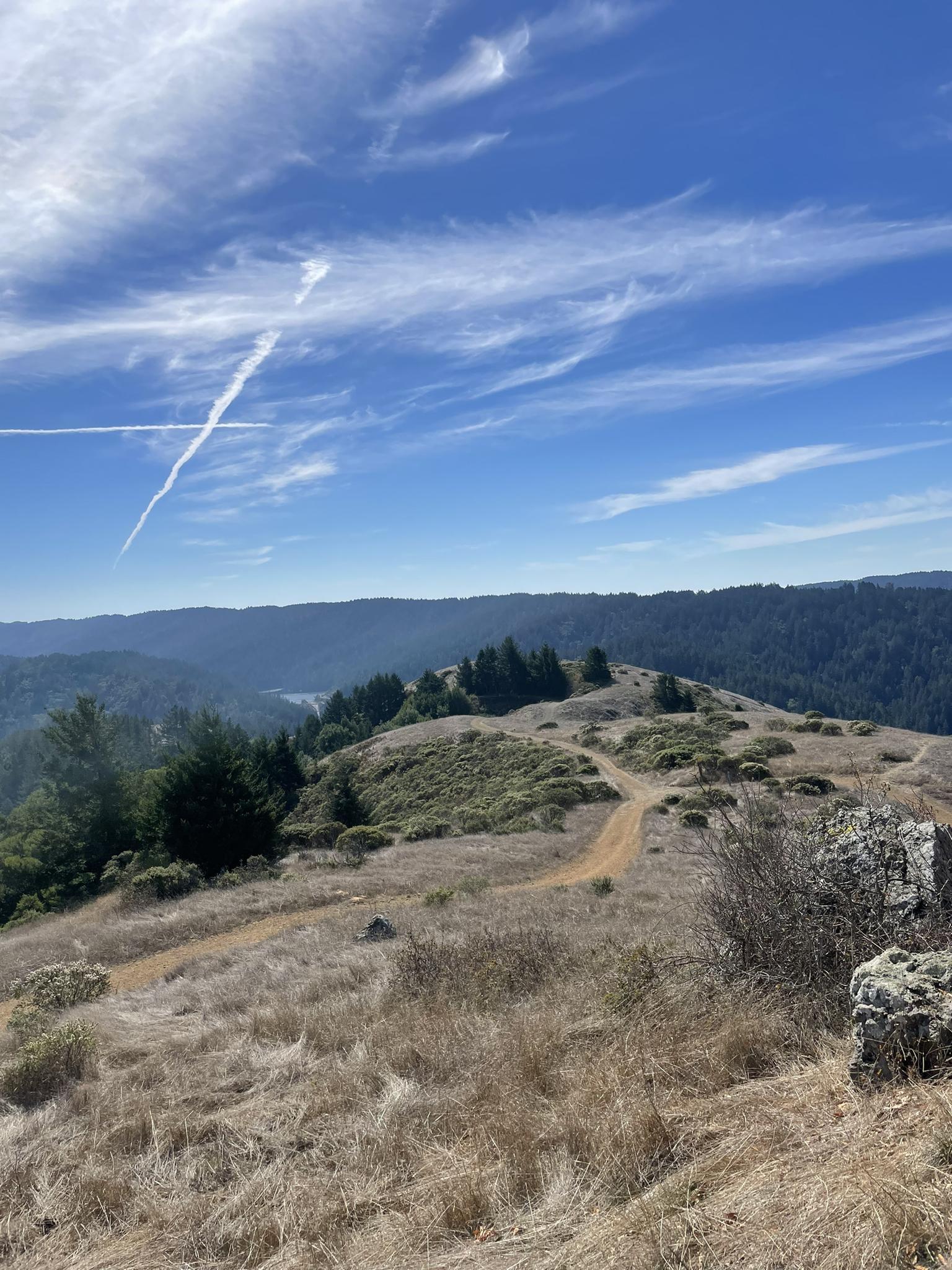 A dirt trail winds through dry grassy hills under a bright blue sky.
