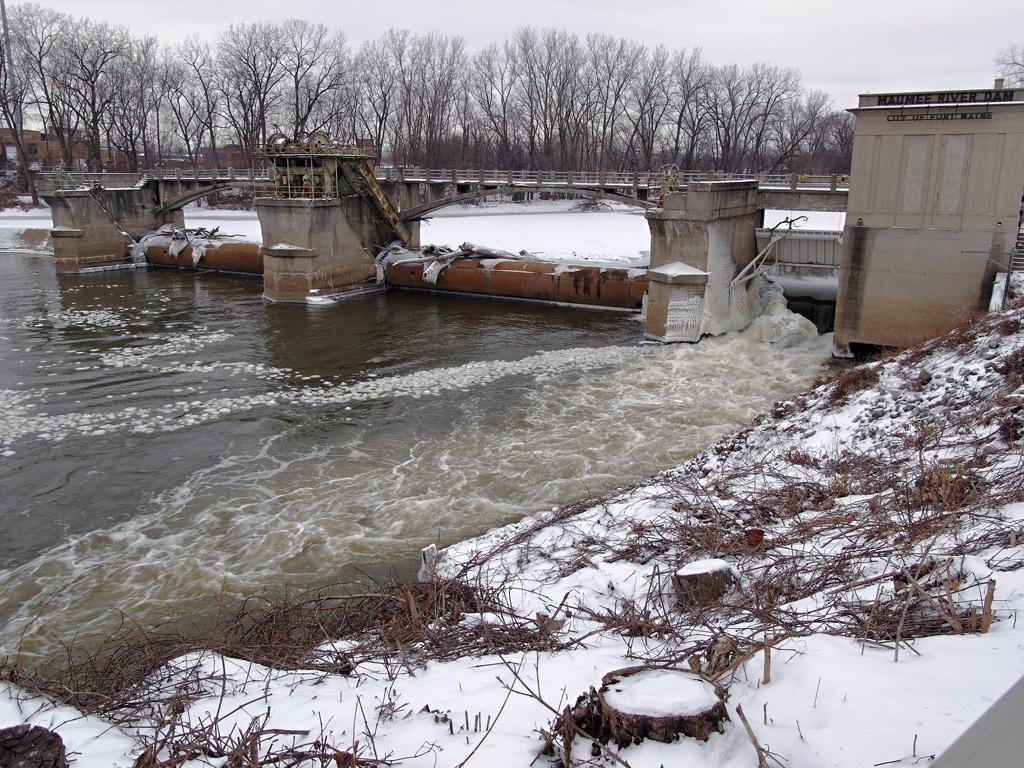 Winter scene with a dam and bridge over a partially frozen, flowing river.