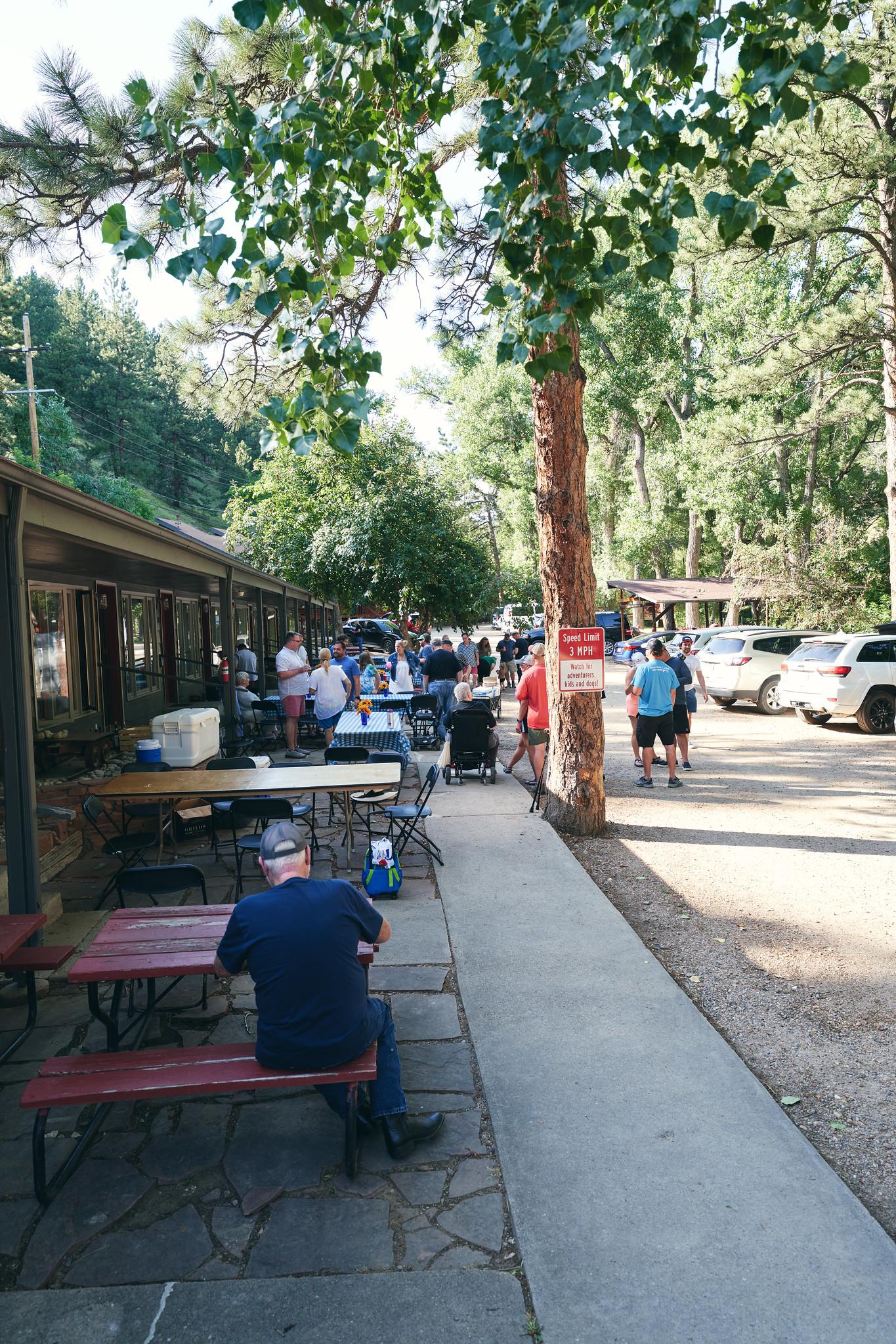 People gather at outdoor tables next to a building, with cars and trees.