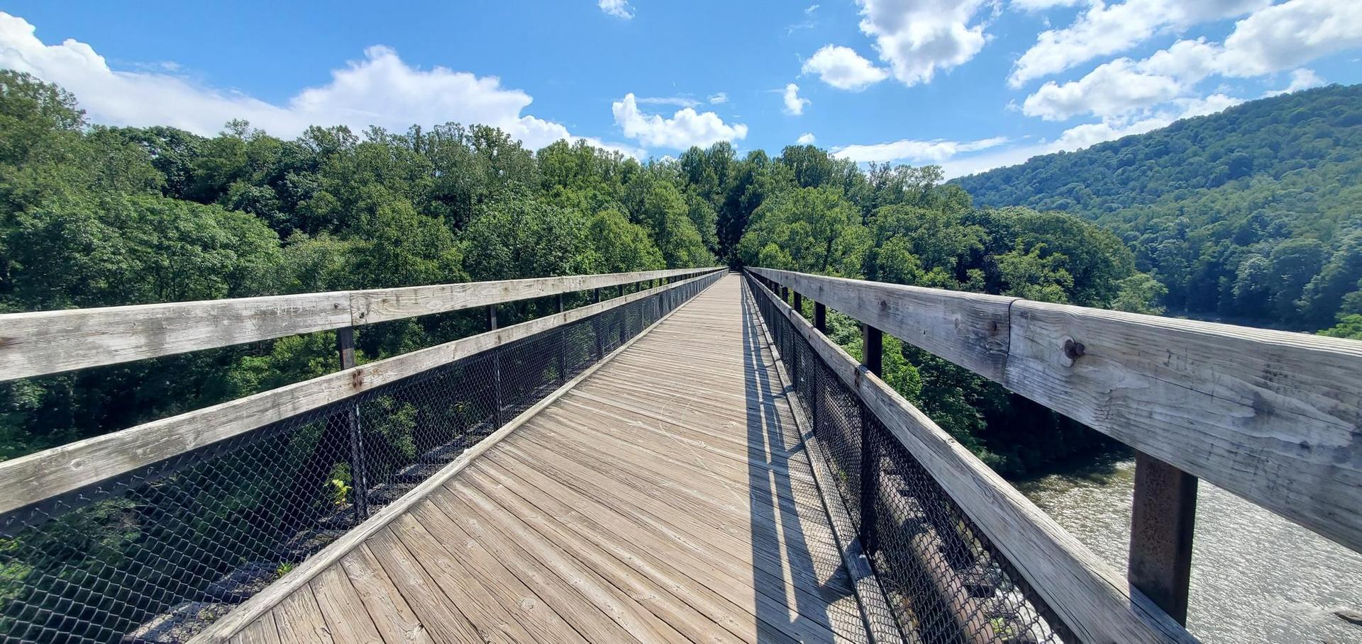 Long wooden bridge over a river, surrounded by green trees and blue sky.