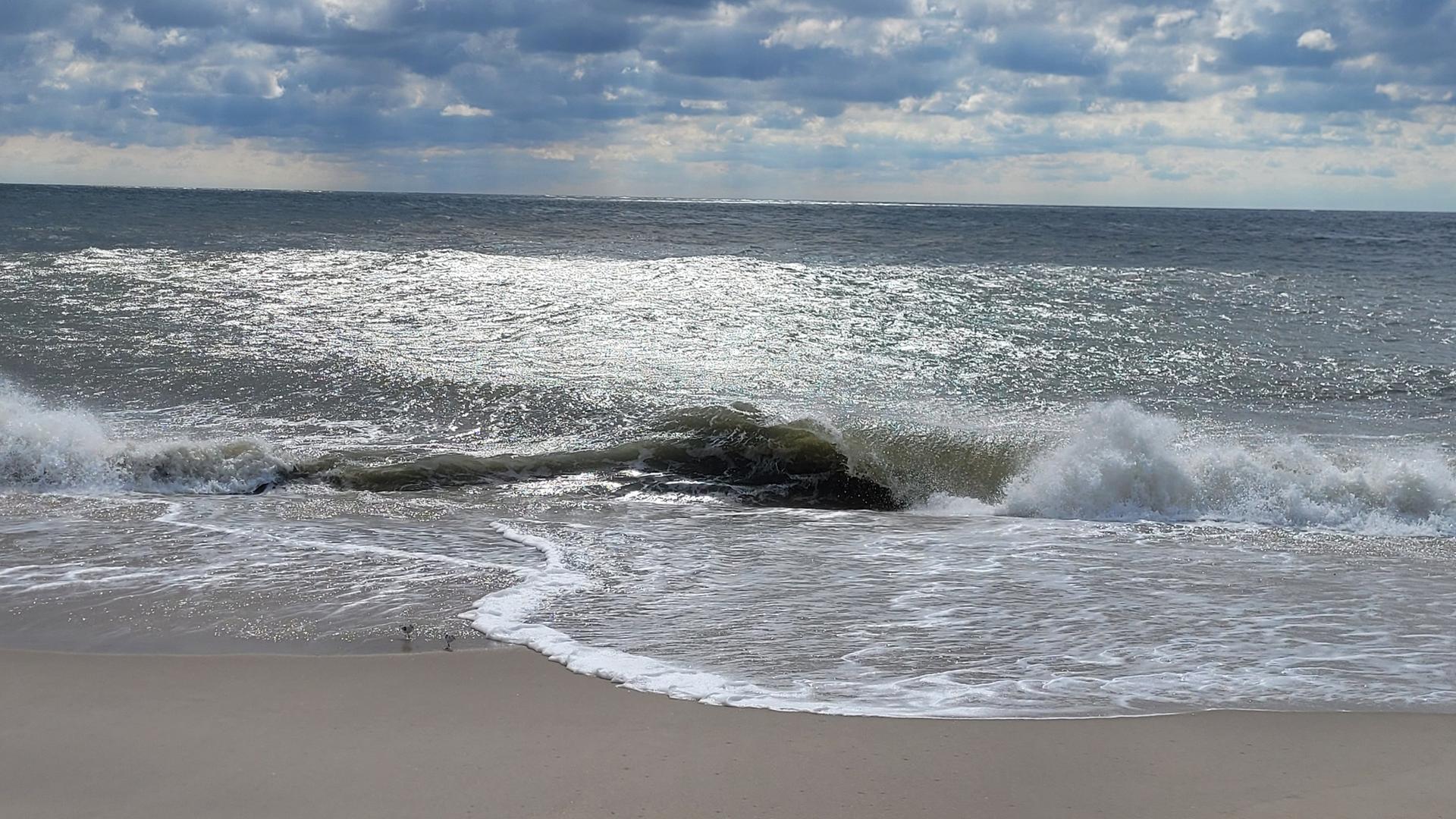 Waves crash on a sandy beach under a cloudy sky, sun glinting.