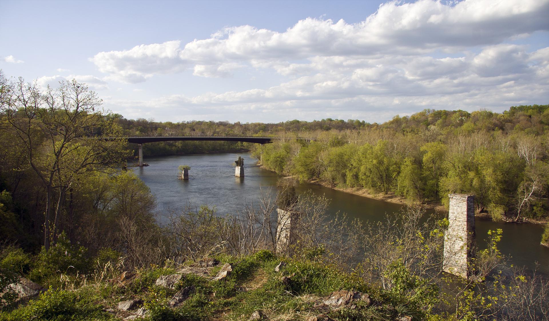 Scenic river valley with a bridge, lush trees, and cloudy daytime sky.