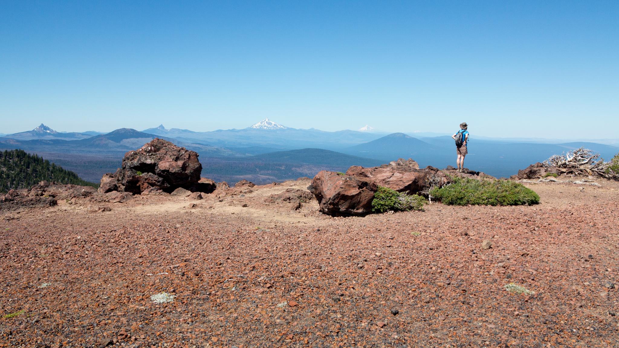 Hiker stands on a rocky summit, admiring distant mountains under clear skies.