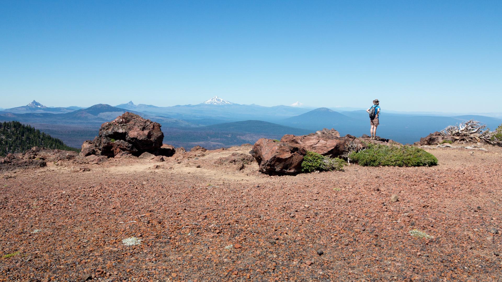 Hiker stands on a rocky summit, admiring distant mountains under clear skies.