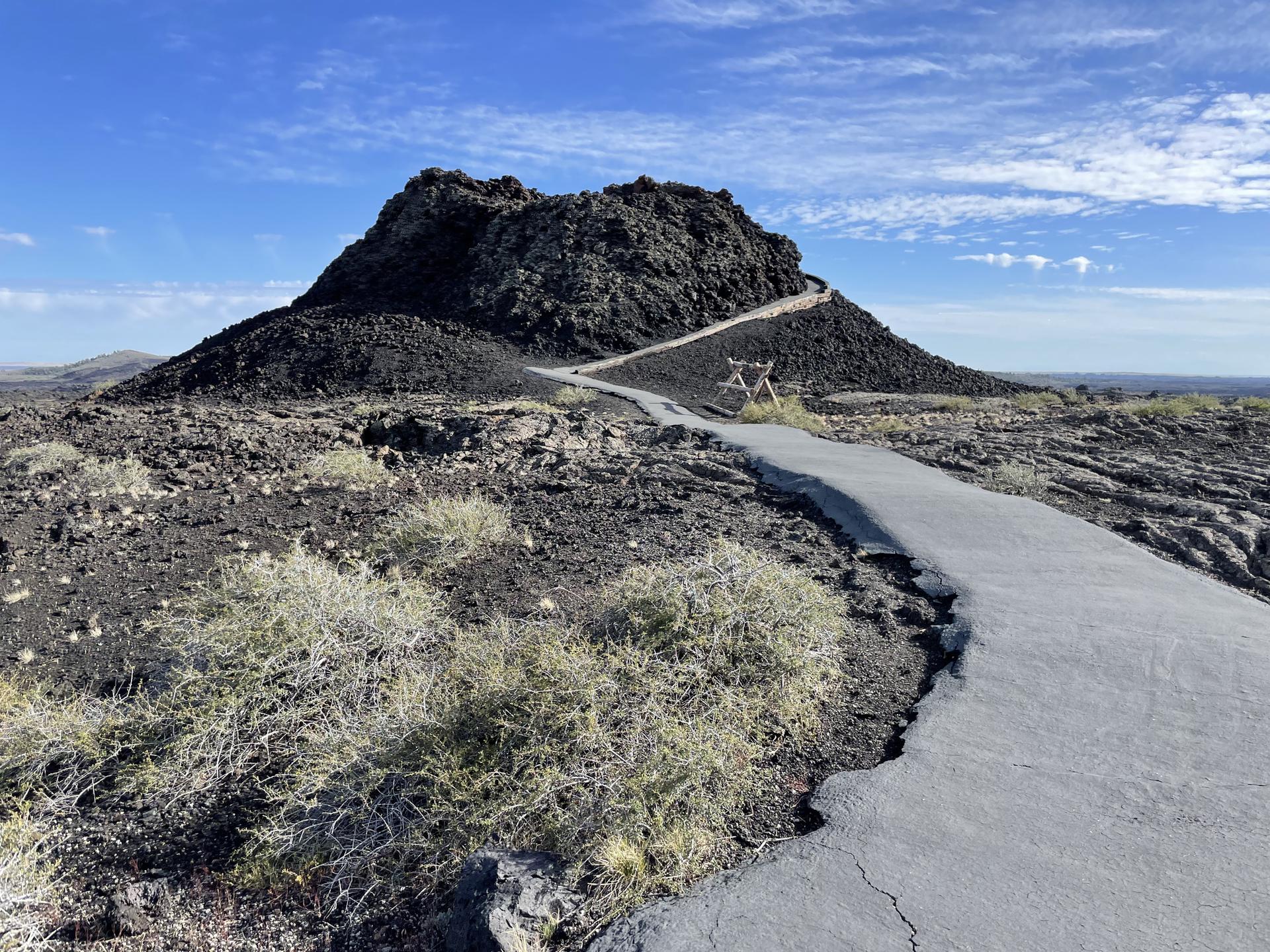 Paved path ascends a dark volcanic hill with sparse vegetation under a bright sky.