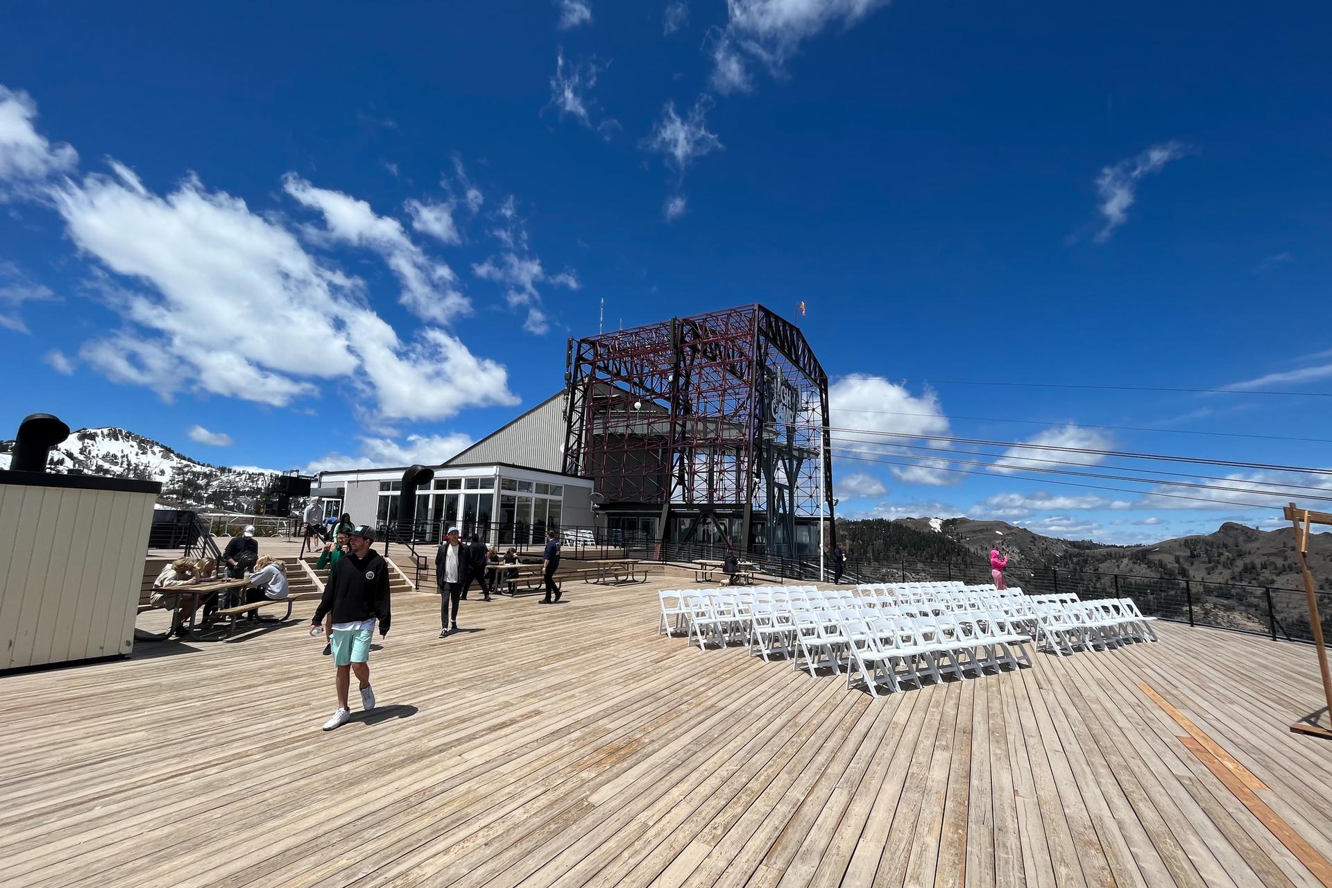 People on a large wooden deck with white chairs and a modern building under blue sky.