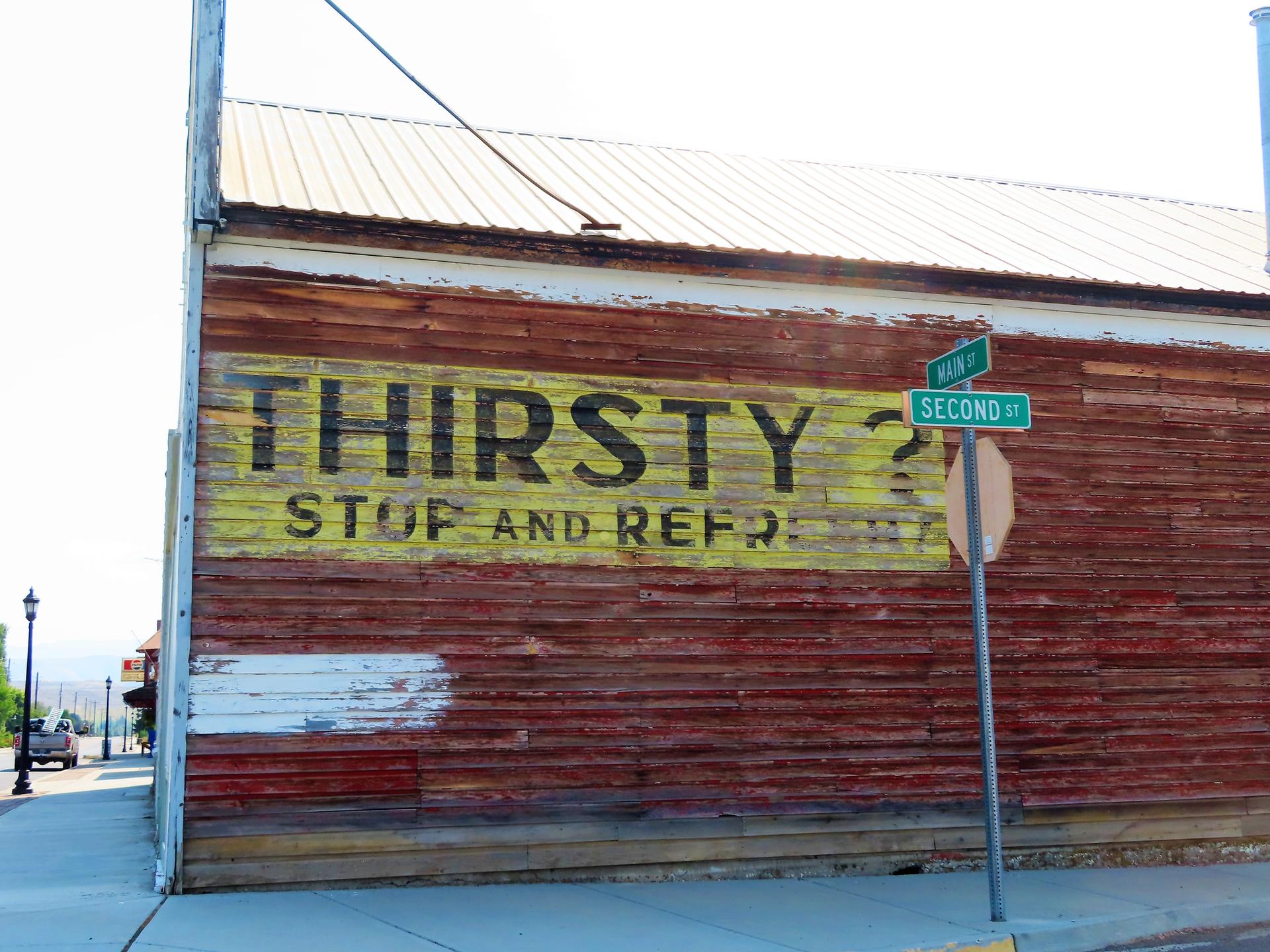 Weathered wooden building with "THIRSTY?" painted sign, street signs, and bright daylight.