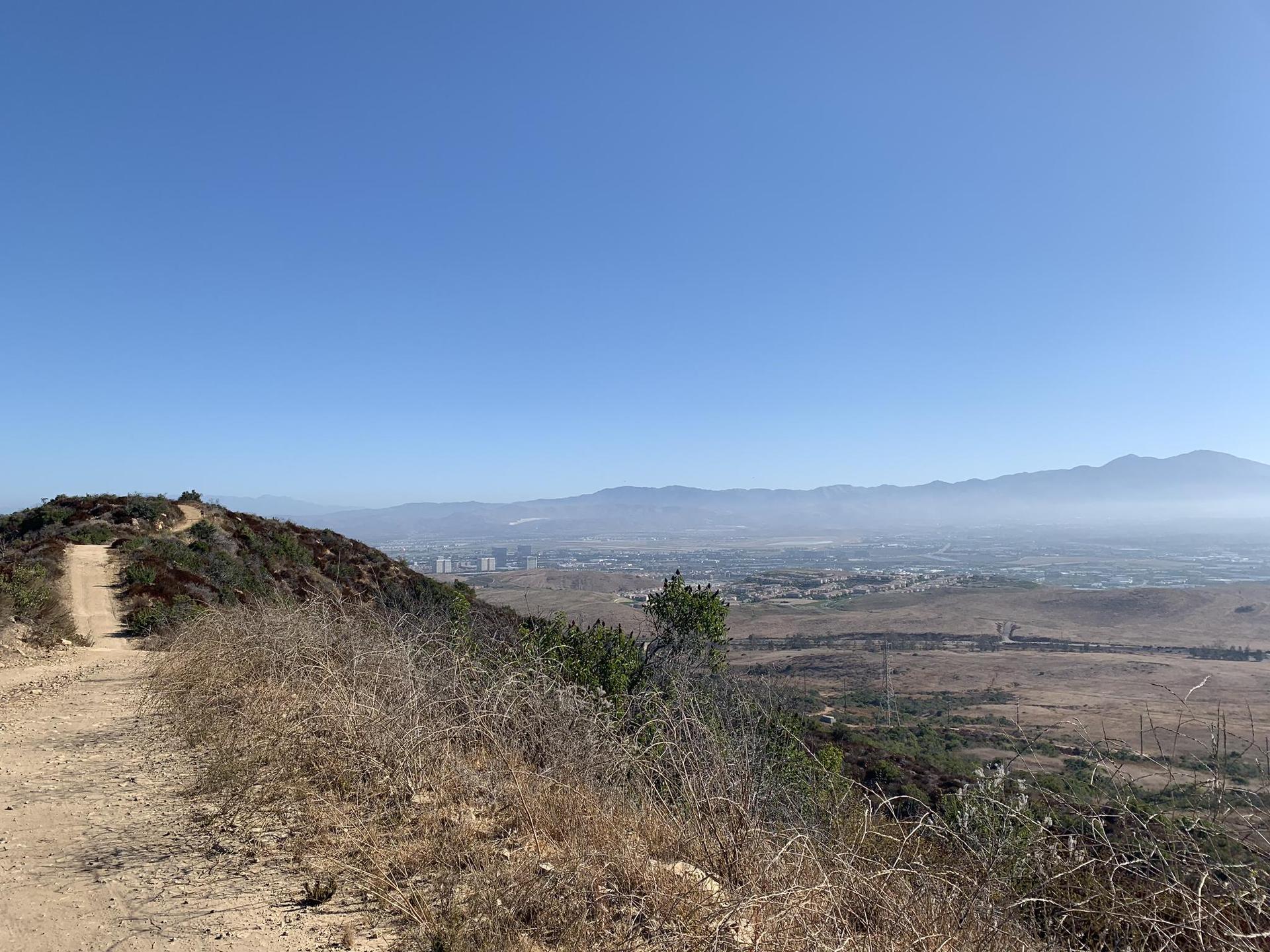 Dirt trail on a dry hill overlooking a hazy valley and distant mountains.
