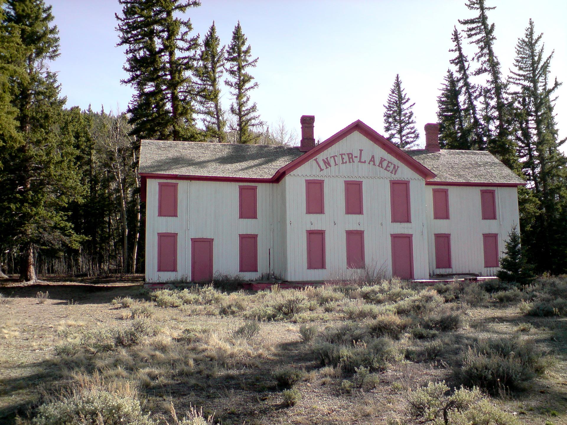 An old white building with red trim stands in a clearing surrounded by pine trees.