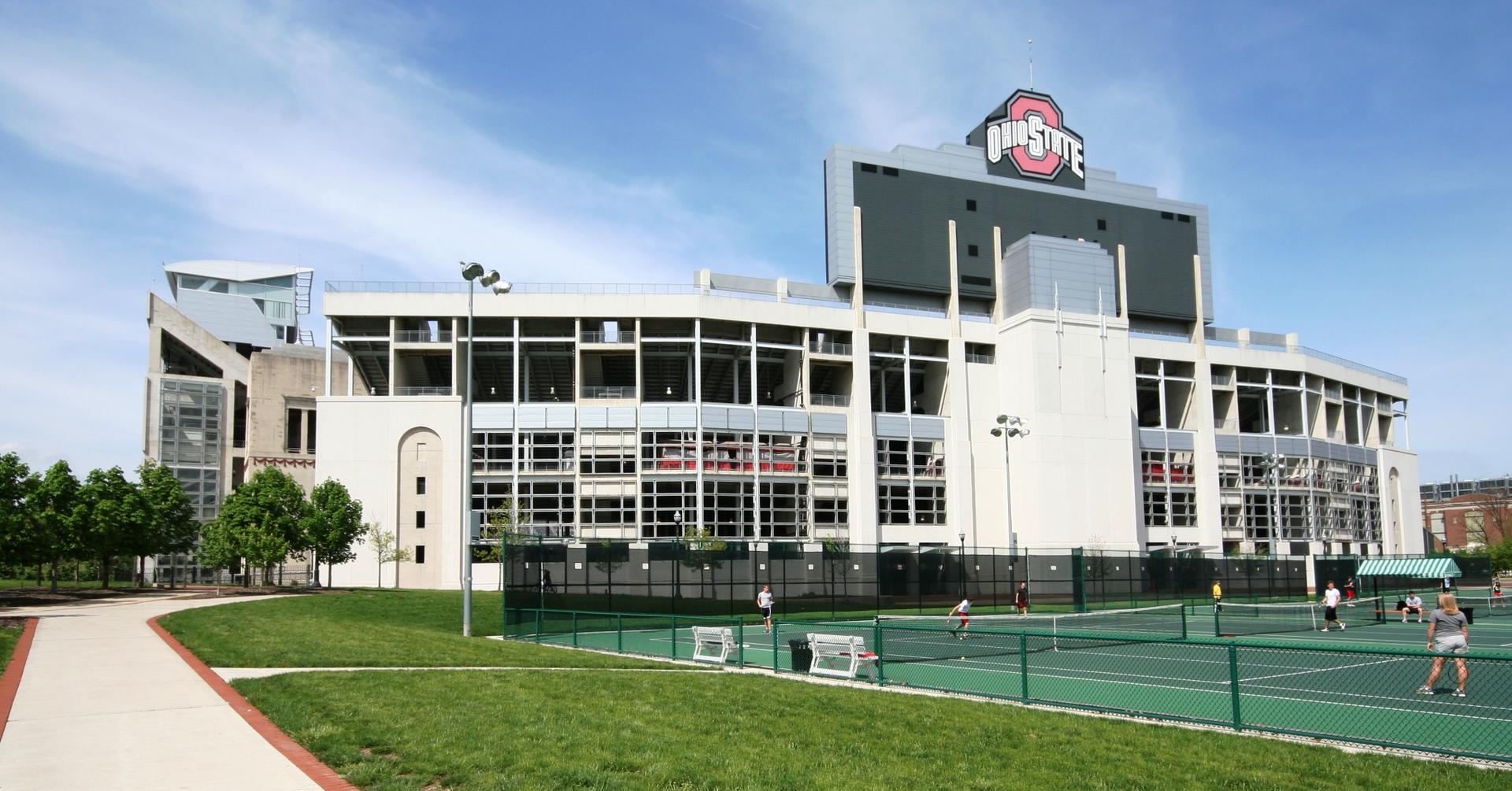 Large stadium overlooks tennis courts with people playing under a clear sky.