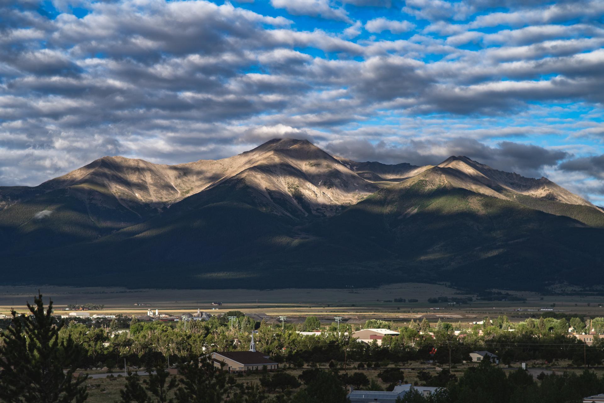 Dramatic cloud shadows fall across a large mountain range above a distant town.