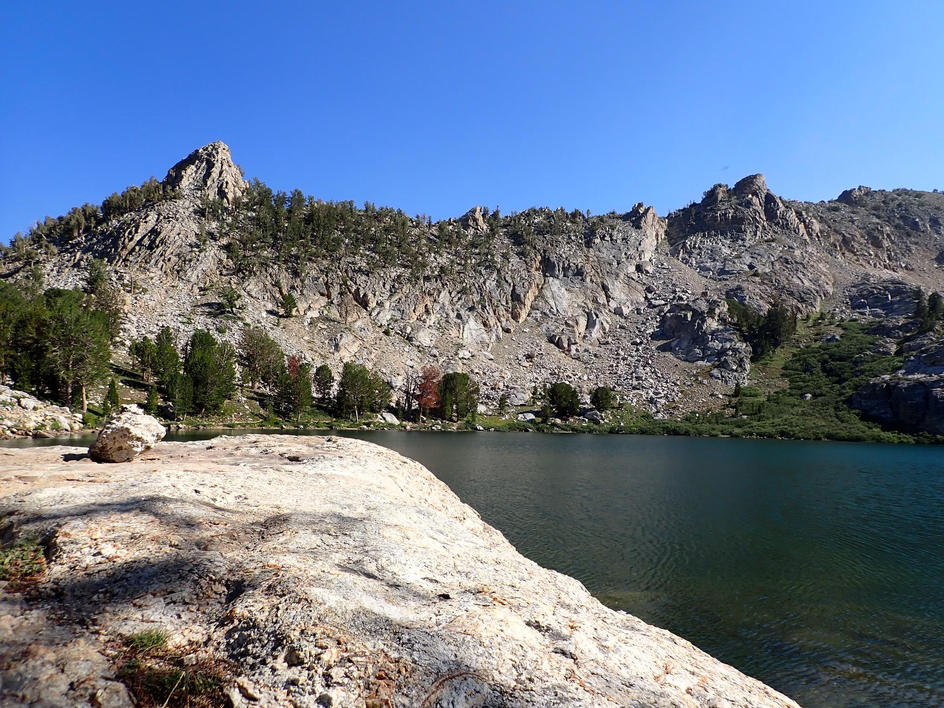 Bright mountain lake with rugged rocky peaks and scattered trees under clear sky.
