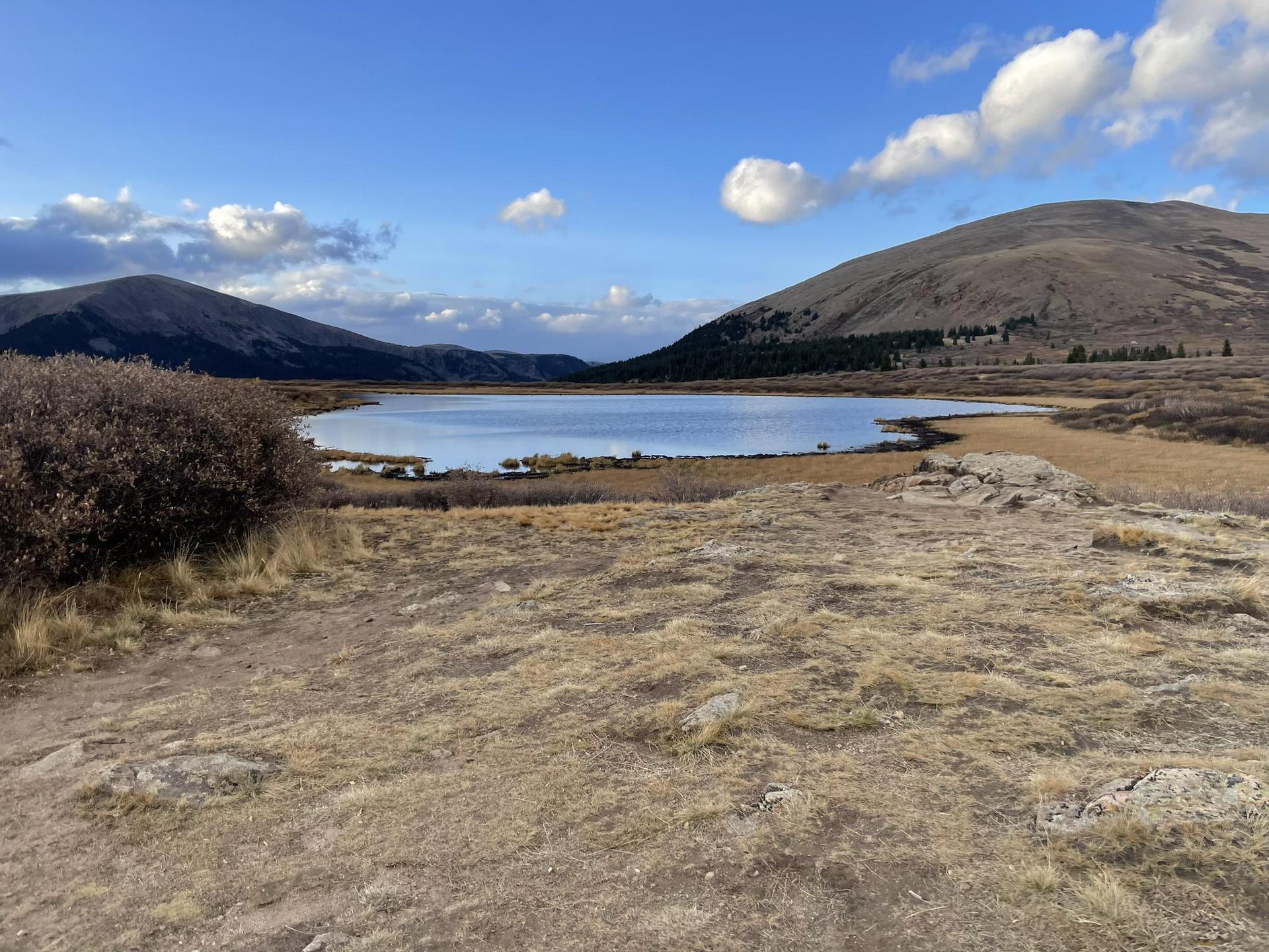 A calm lake surrounded by dry grass and mountains under a blue sky.