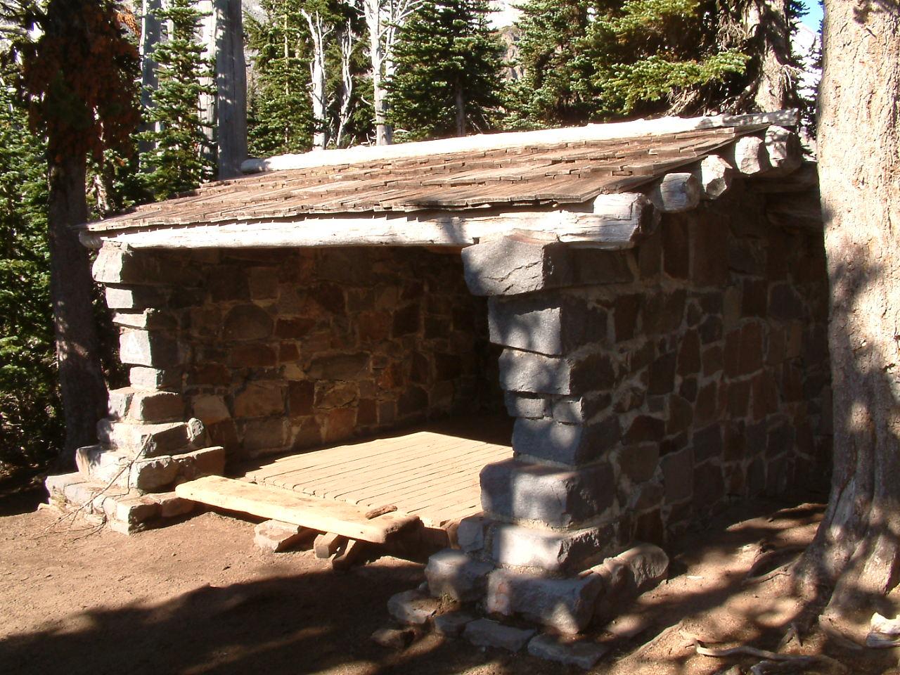 A rustic stone shelter with a wooden floor and roof in a sunny forest.
