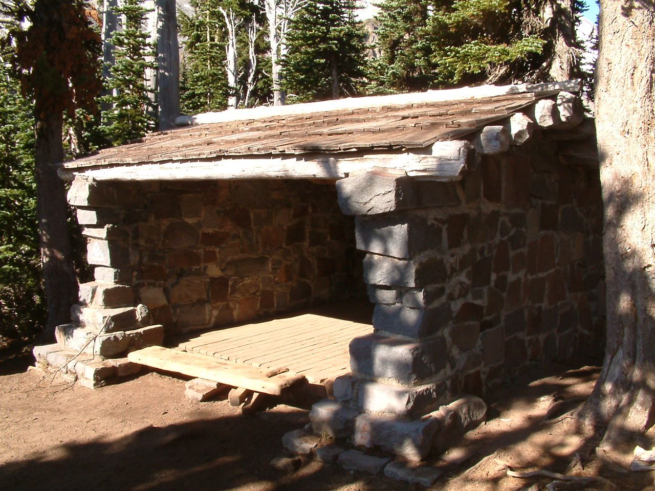 A rustic stone shelter with a wooden floor and roof in a sunny forest. - Summerland Trail