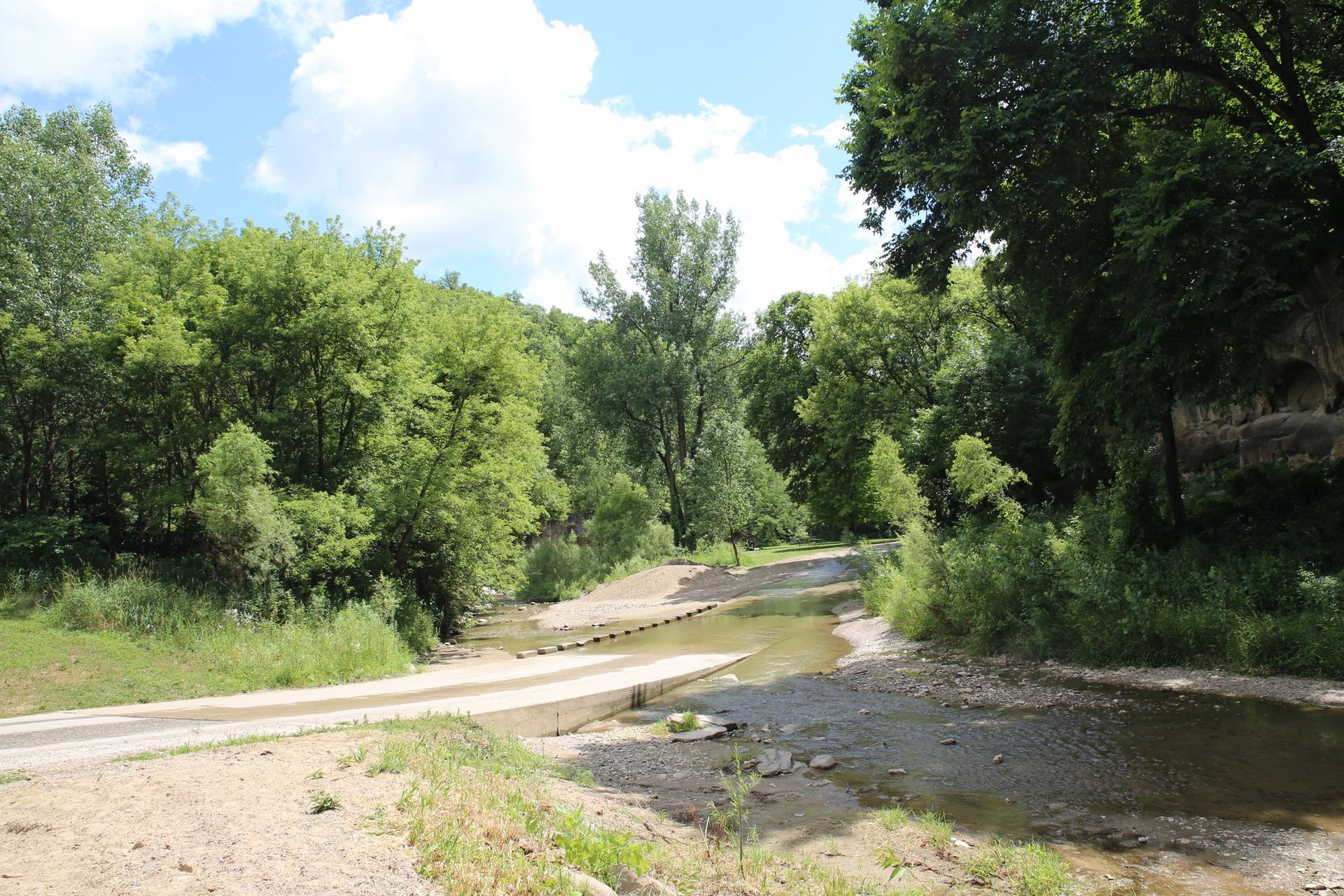 A concrete road crosses a shallow stream surrounded by dense green trees on a sunny day.