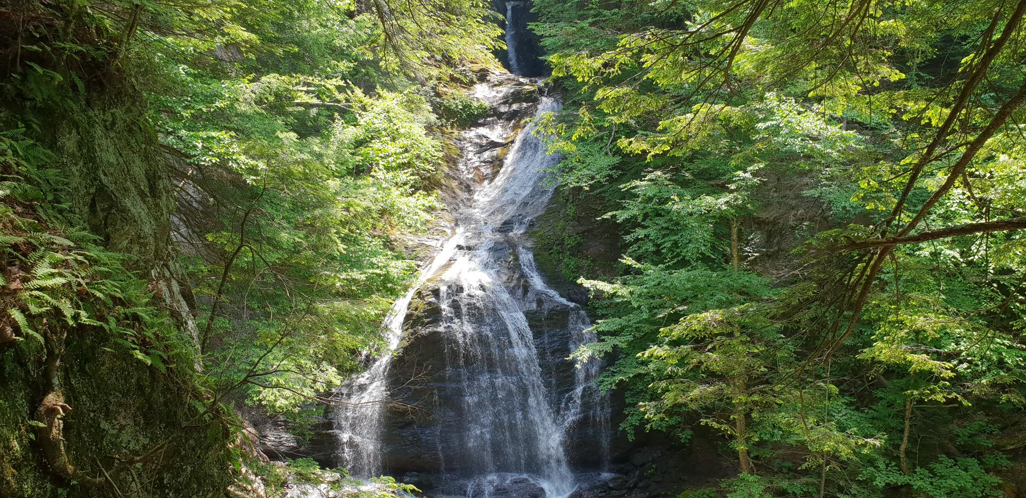 A tall waterfall cascades down a rocky cliff in a lush, sunlit forest.