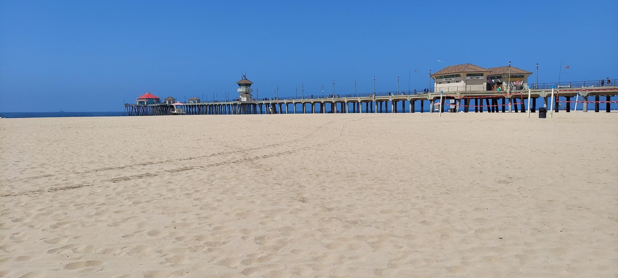 Long pier extending into the ocean from a wide sandy beach under clear blue sky.