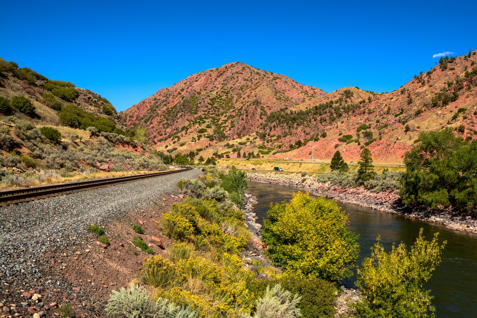Sunny landscape with railway, river, and red-rock mountains under blue sky.