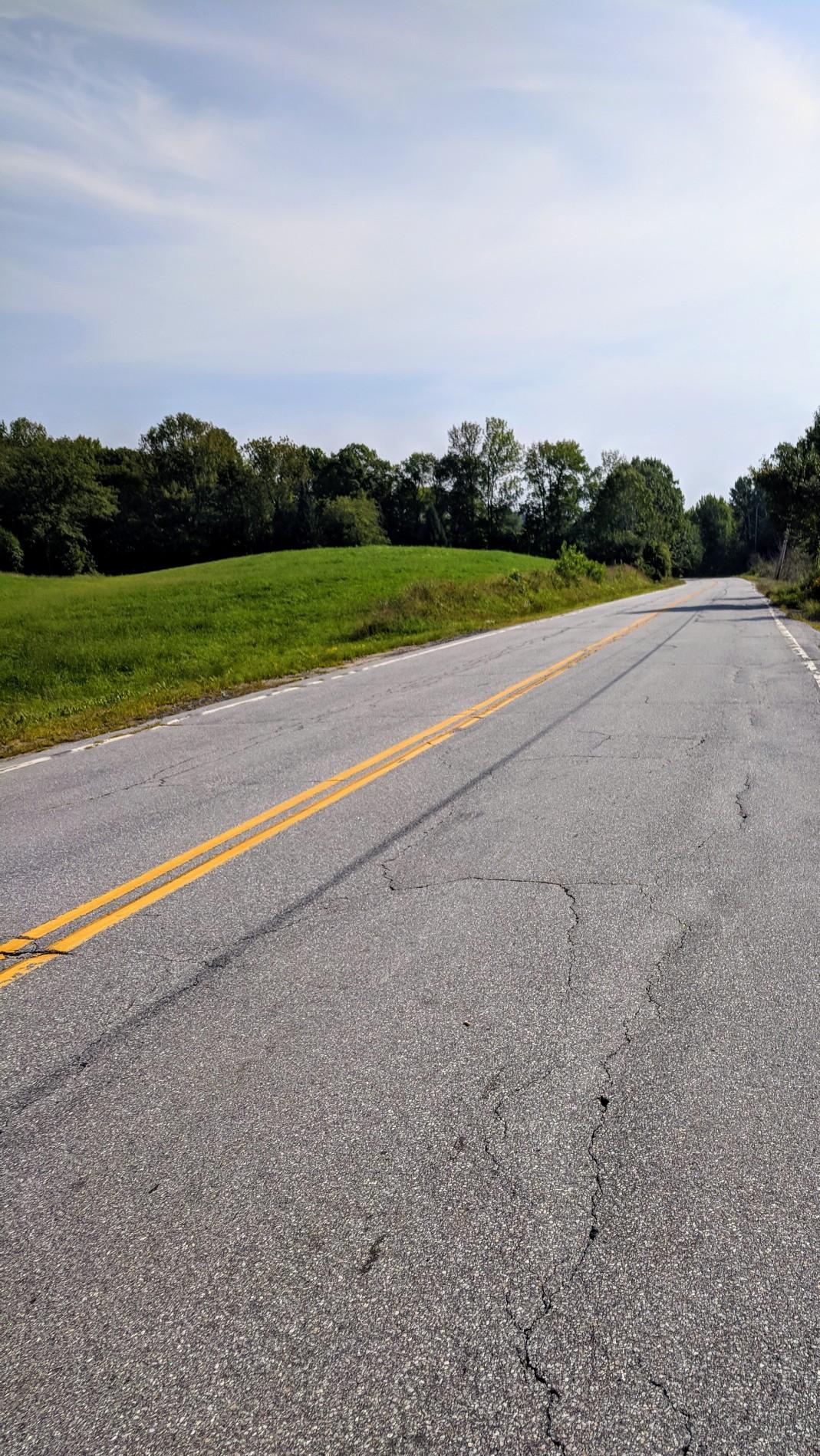 Empty paved road with double yellow lines, green hills, and trees under daylight.