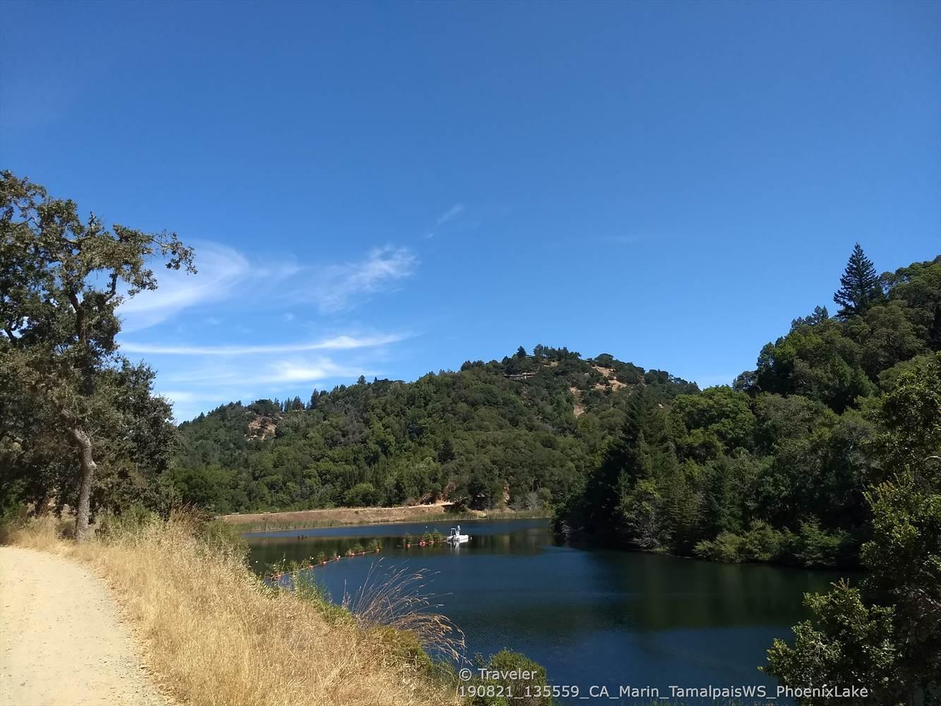 A dirt path beside a lake with forested hills under a bright blue sky.
