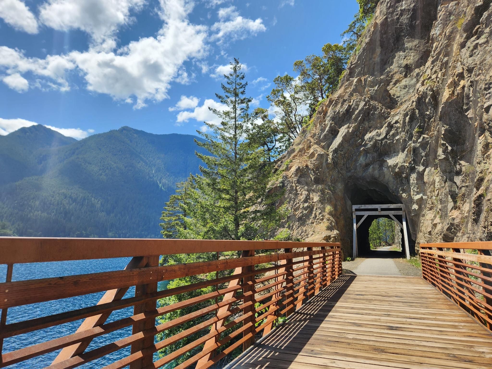 Wooden boardwalk next to a lake, leading to a tunnel through a rocky cliff. - Spruce Railroad Trail