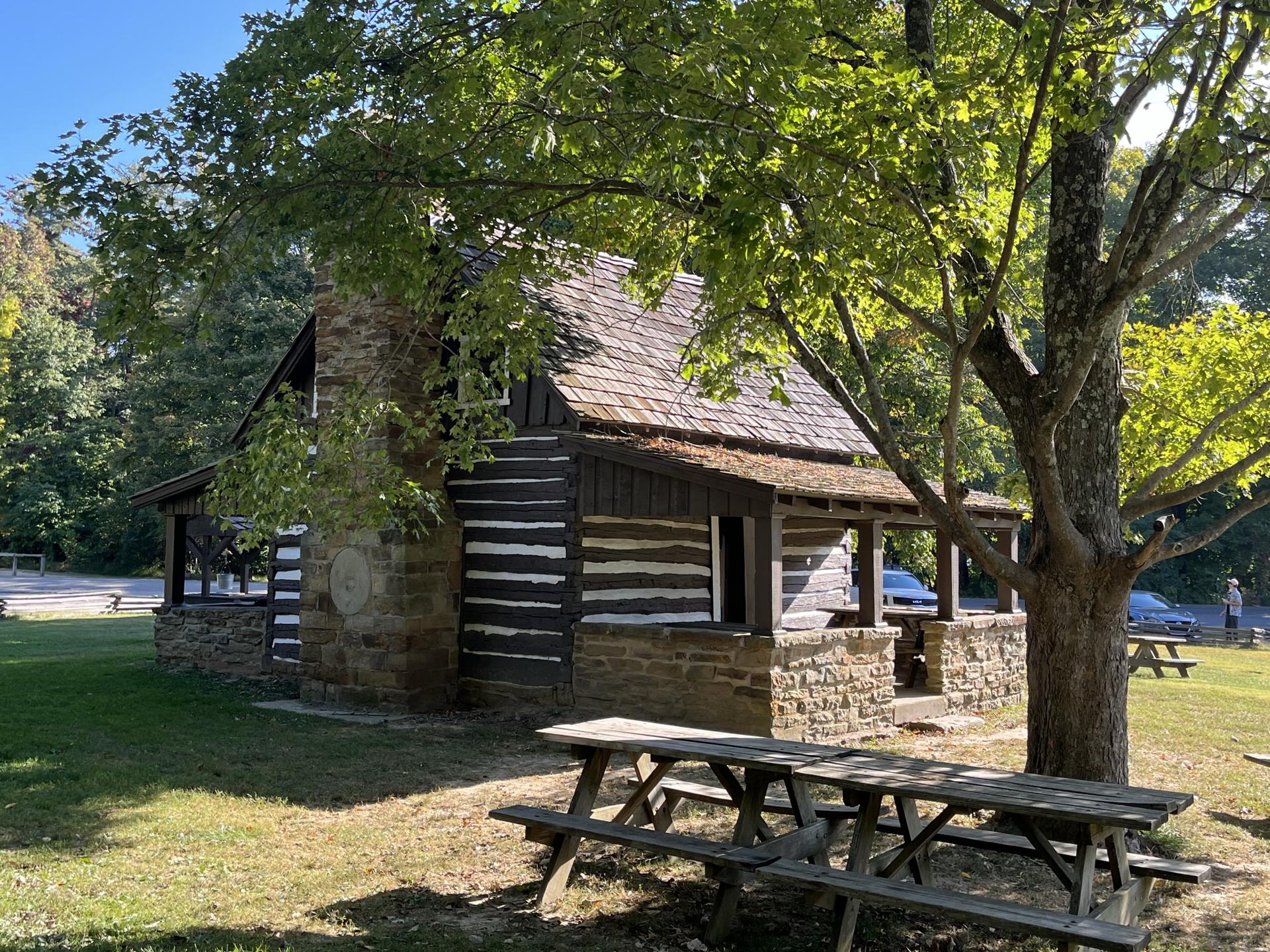 Rustic log cabin with stone chimney, green trees, and picnic tables on a sunny day.