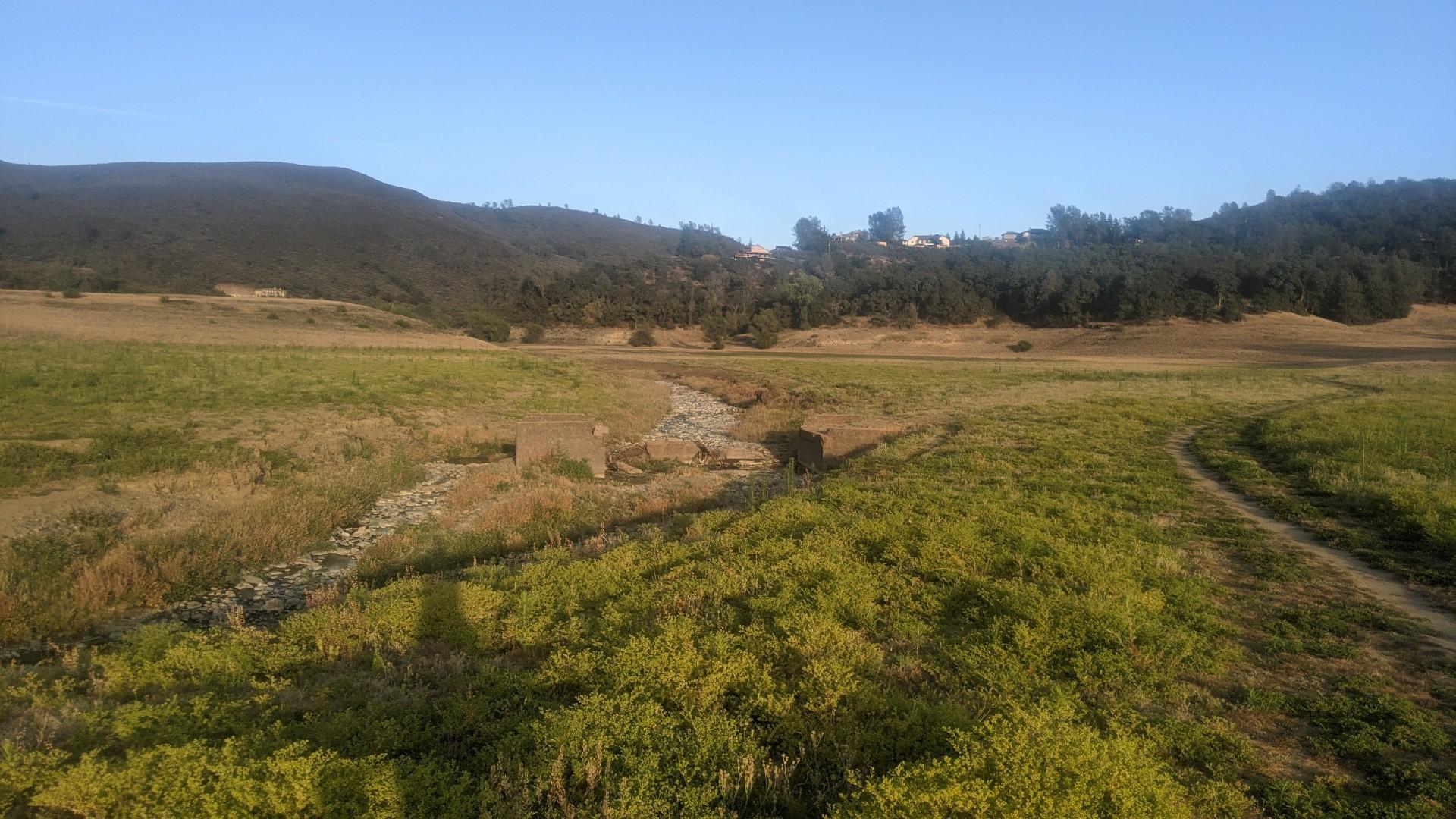 Wide grassy field with a dirt path leading towards distant tree-covered hills.