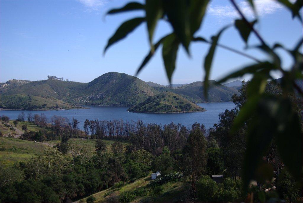 Blue lake surrounded by green hills, framed by blurred foreground leaves.