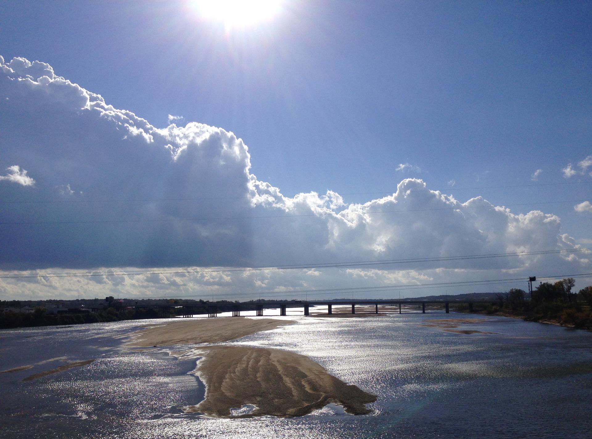 Bright sun over a wide river with a sandy island and distant bridge.