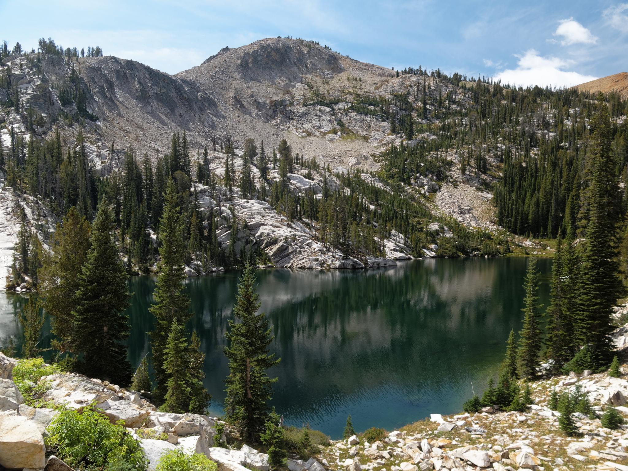 Calm mountain lake reflecting pine trees and rocky slopes under a bright sky.